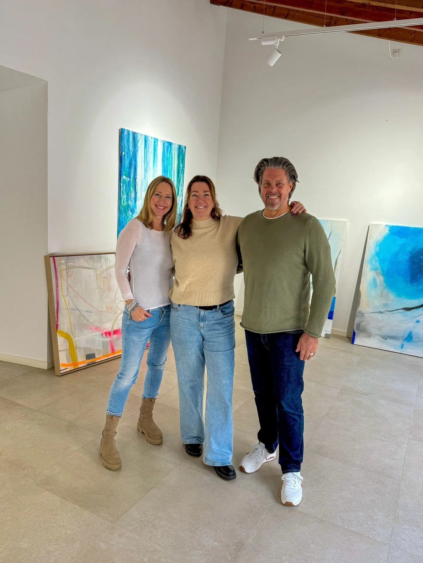 Three smiling people posing together in an art gallery with colorful abstract paintings in the background. The woman on the left is wearing a white long-sleeve shirt, light blue jeans, and tan boots. The woman in the middle is wearing a beige sweater and blue jeans. The man on the right is wearing a green sweater, dark blue jeans, and white sneakers.