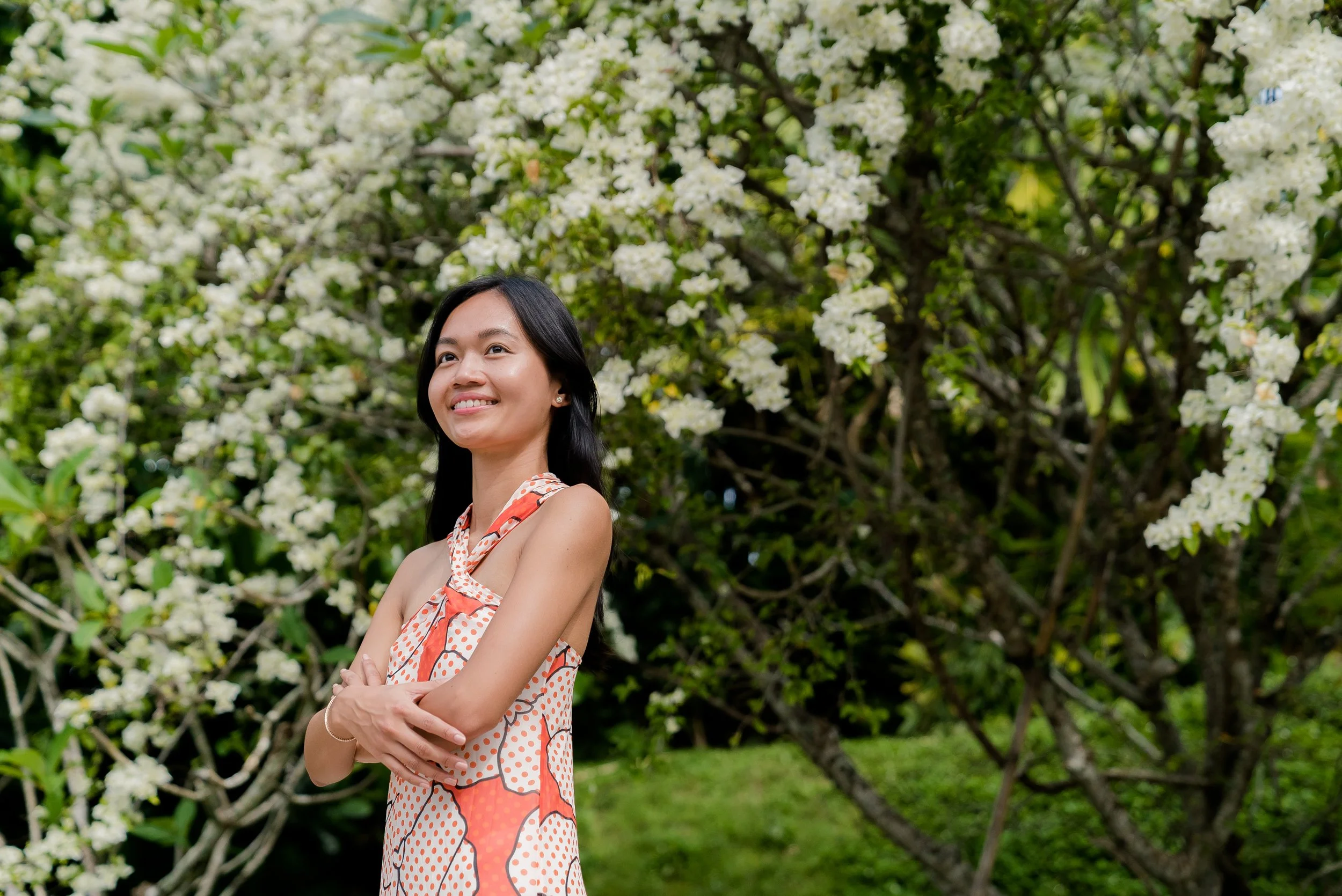 A woman with long dark hair smiling and standing outdoors with crossed arms, in front of a flowering tree with white blossoms.