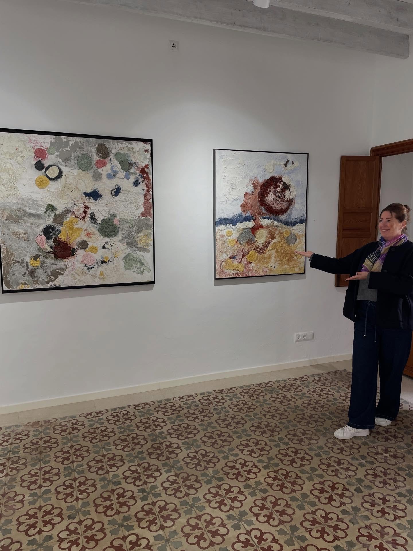 A woman standing in an art gallery, smiling, and gesturing towards two abstract paintings on a white wall. The gallery has patterned floor tiles and a wooden door on the right side.