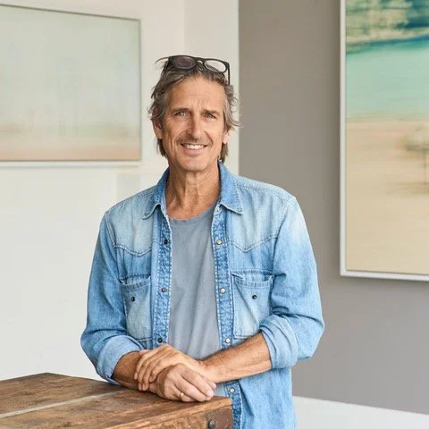 Man with glasses on head, smiling, wearing a denim shirt, standing in an art gallery.
