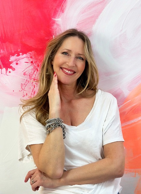 A smiling woman with long blonde hair, wearing a white t-shirt and multiple bracelets, standing in front of an abstract pink and white background.