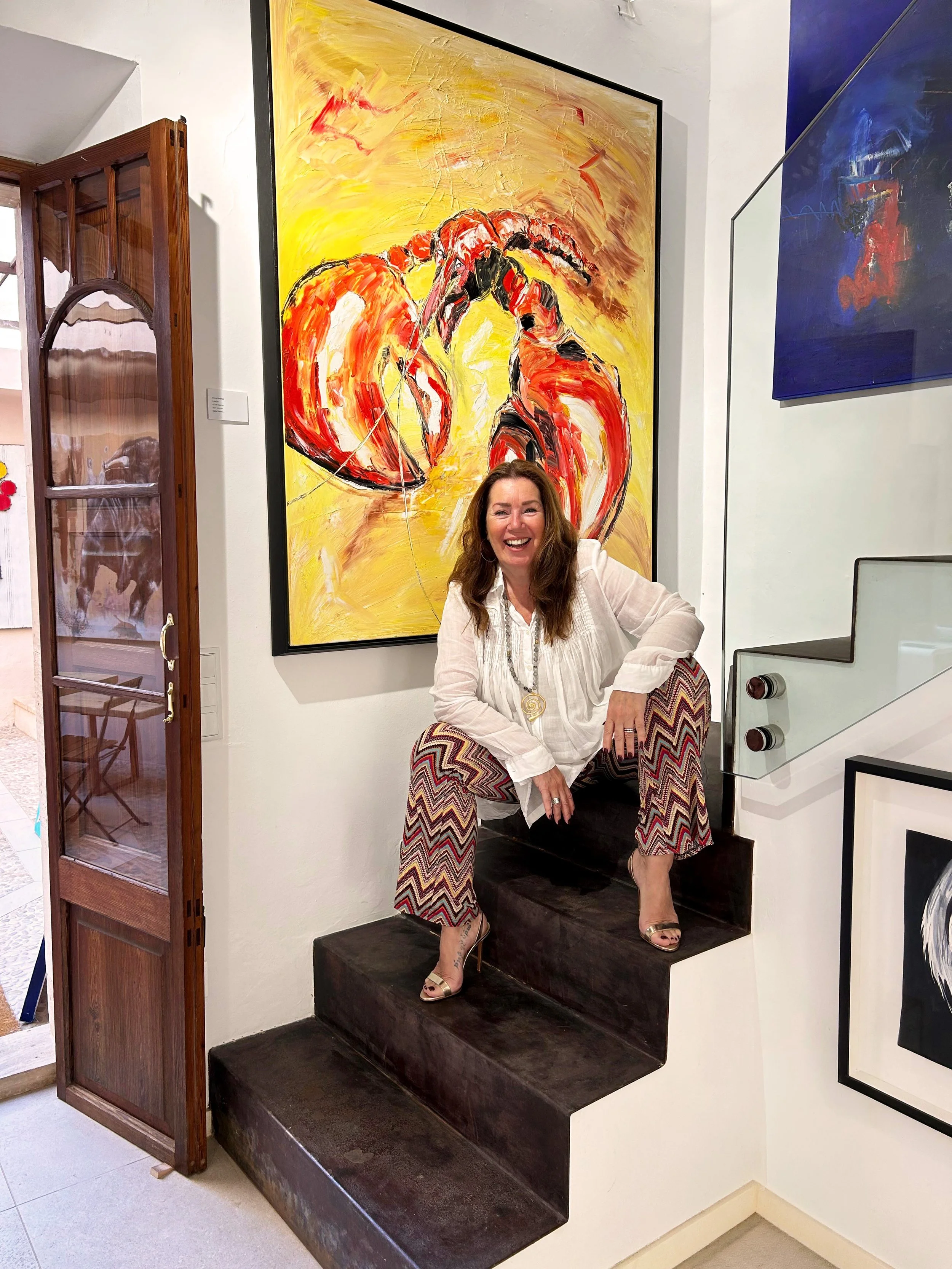 A woman sitting on a dark brown stair in an art gallery, smiling and laughing, with colorful abstract paintings on the white wall behind her.