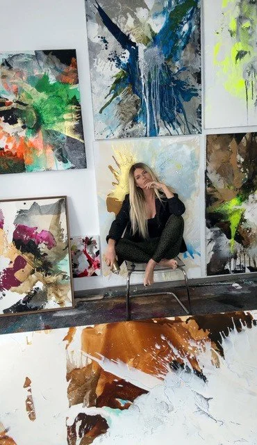 A woman with long blonde hair sitting on a chair in an art studio surrounded by large, colorful abstract paintings on the walls and floor.