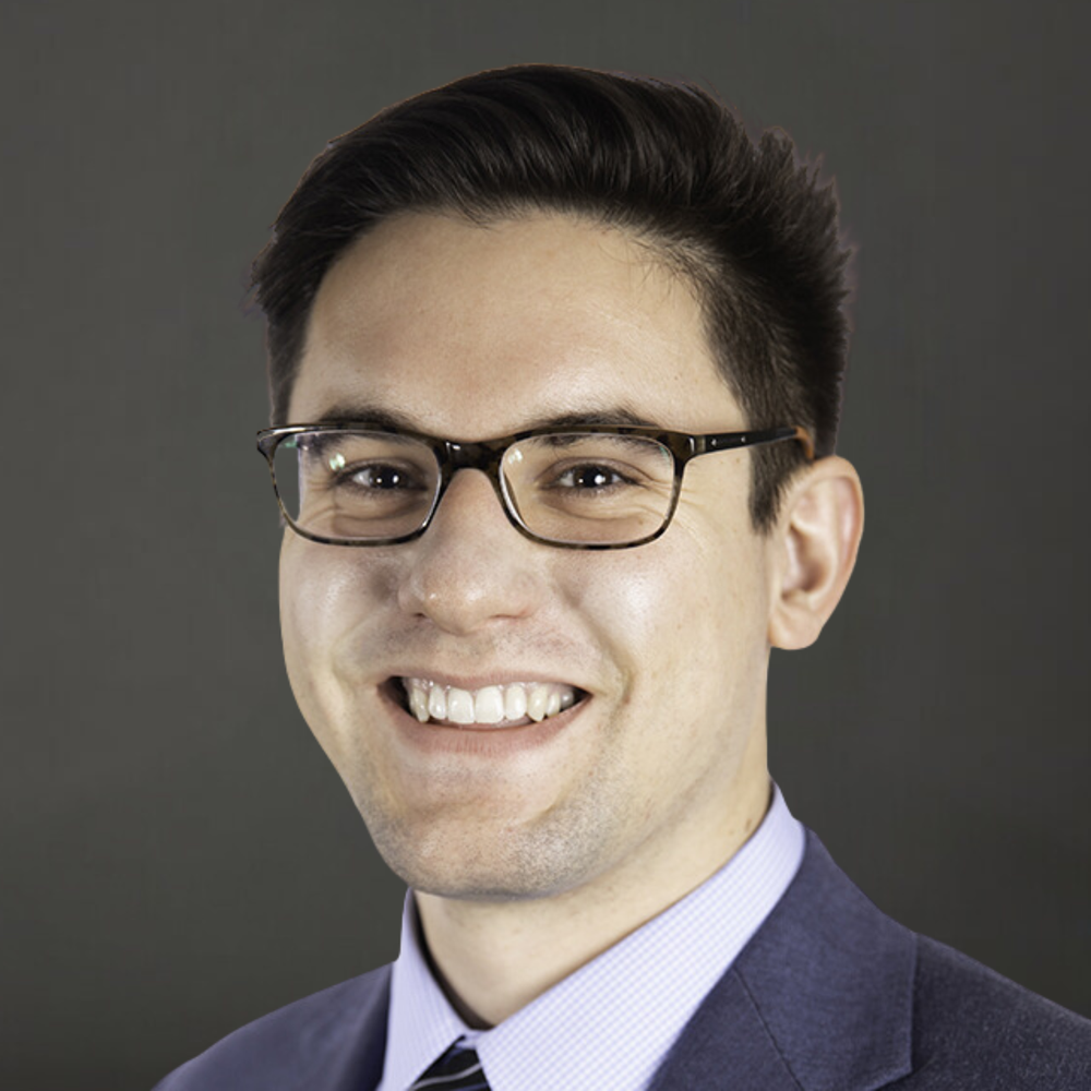 Portrait of a young man with dark hair and glasses, smiling, dressed in a suit and tie against a gray background.