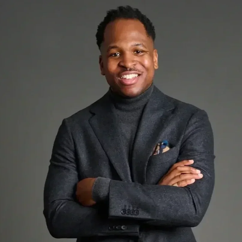 A man in a dark gray blazer and turtleneck smiling, with arms crossed against a gray background.