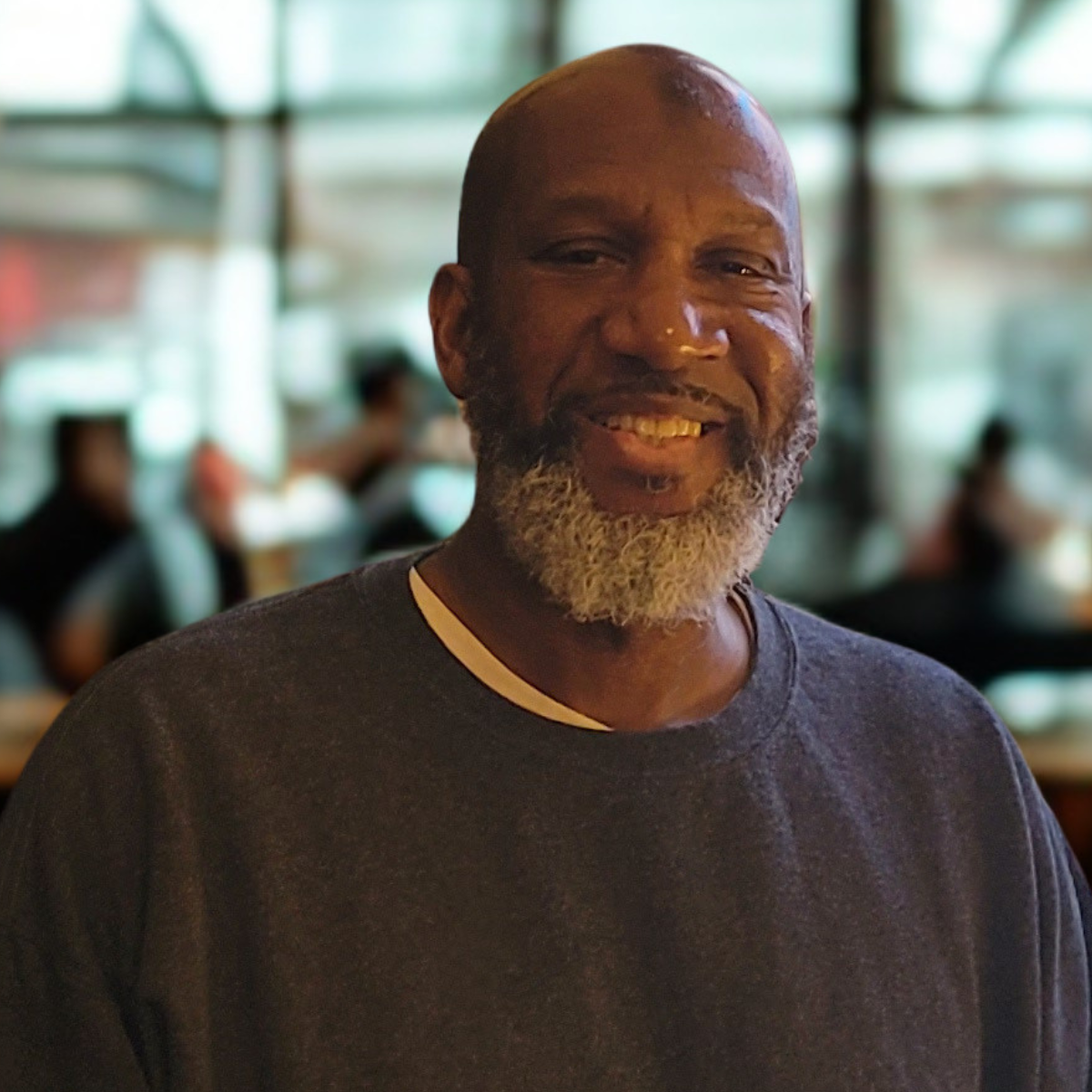 A smiling man with a gray beard wearing a dark t-shirt in a restaurant or cafe, with blurred people and large windows in the background.