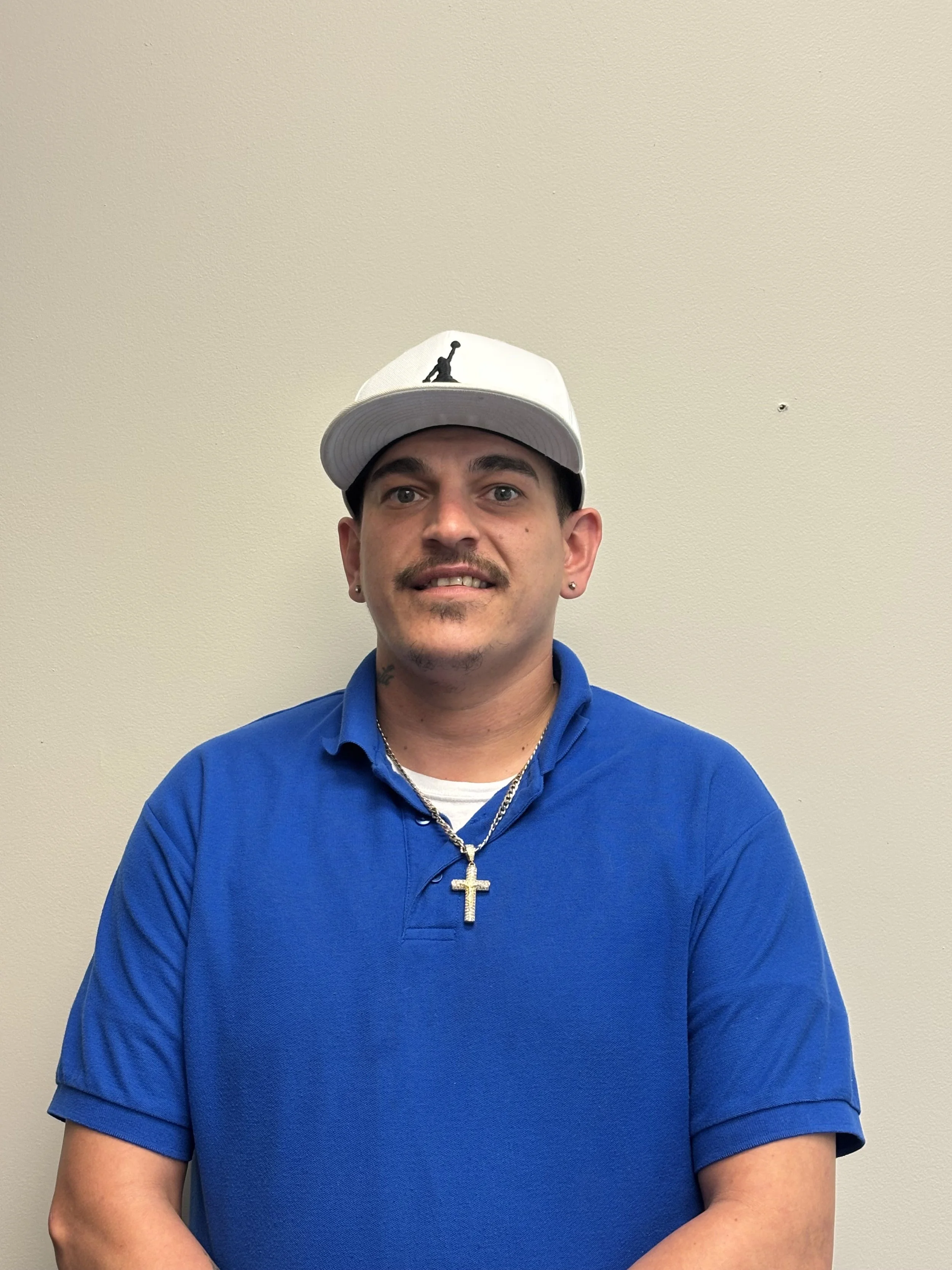 A young man with short dark hair, facial hair, and piercings in both ears wearing a white baseball cap with a black logo, a blue polo shirt, a silver chain necklace with a cross pendant, and standing against a plain beige wall.