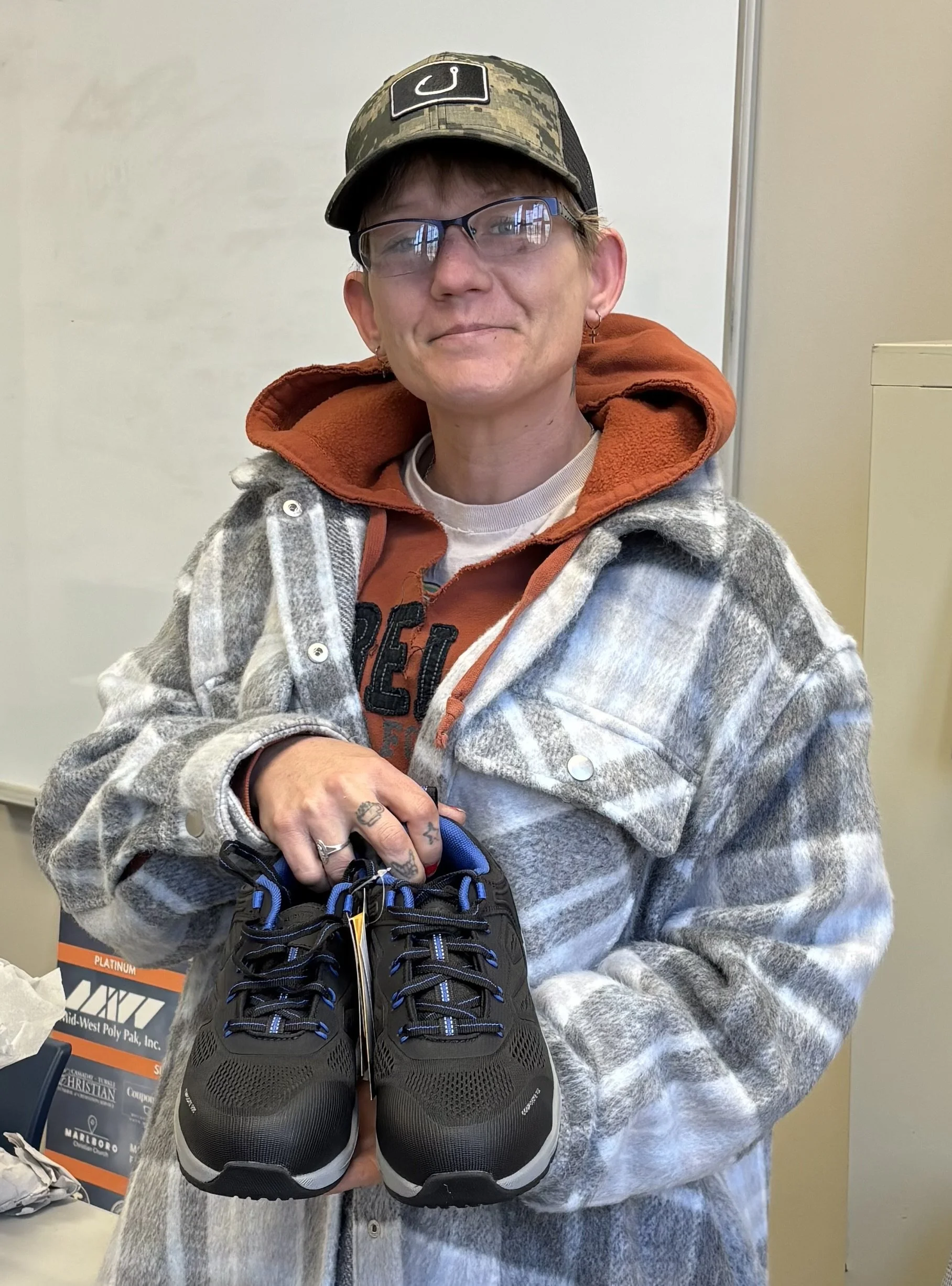 Person with short hair, glasses, and earrings holding a pair of black and gray Nike sneakers in a room with white walls and a whiteboard.