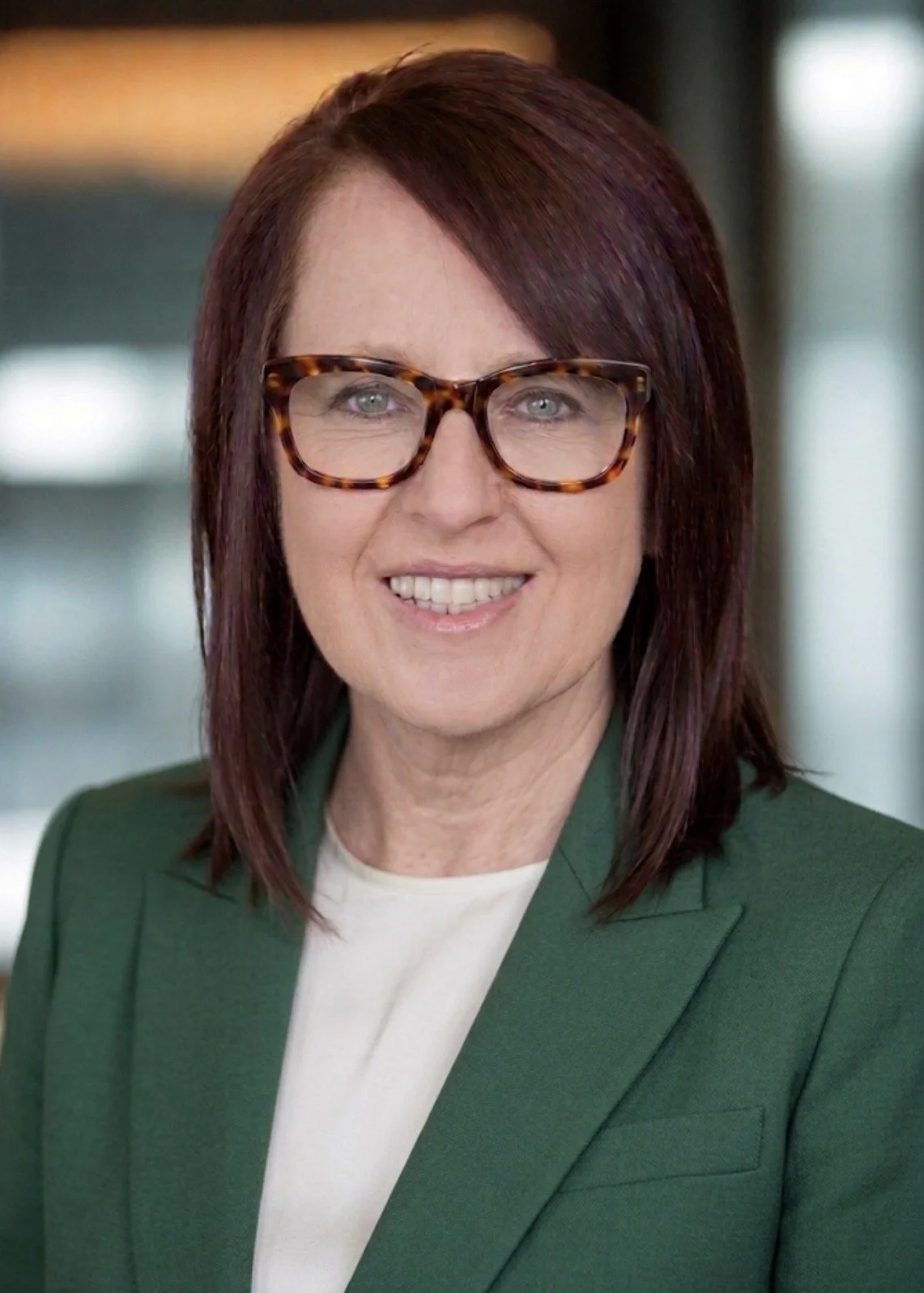 A woman with shoulder-length reddish-brown hair, wearing tortoiseshell glasses, a green blazer, and a white top, smiling in an indoor setting.
