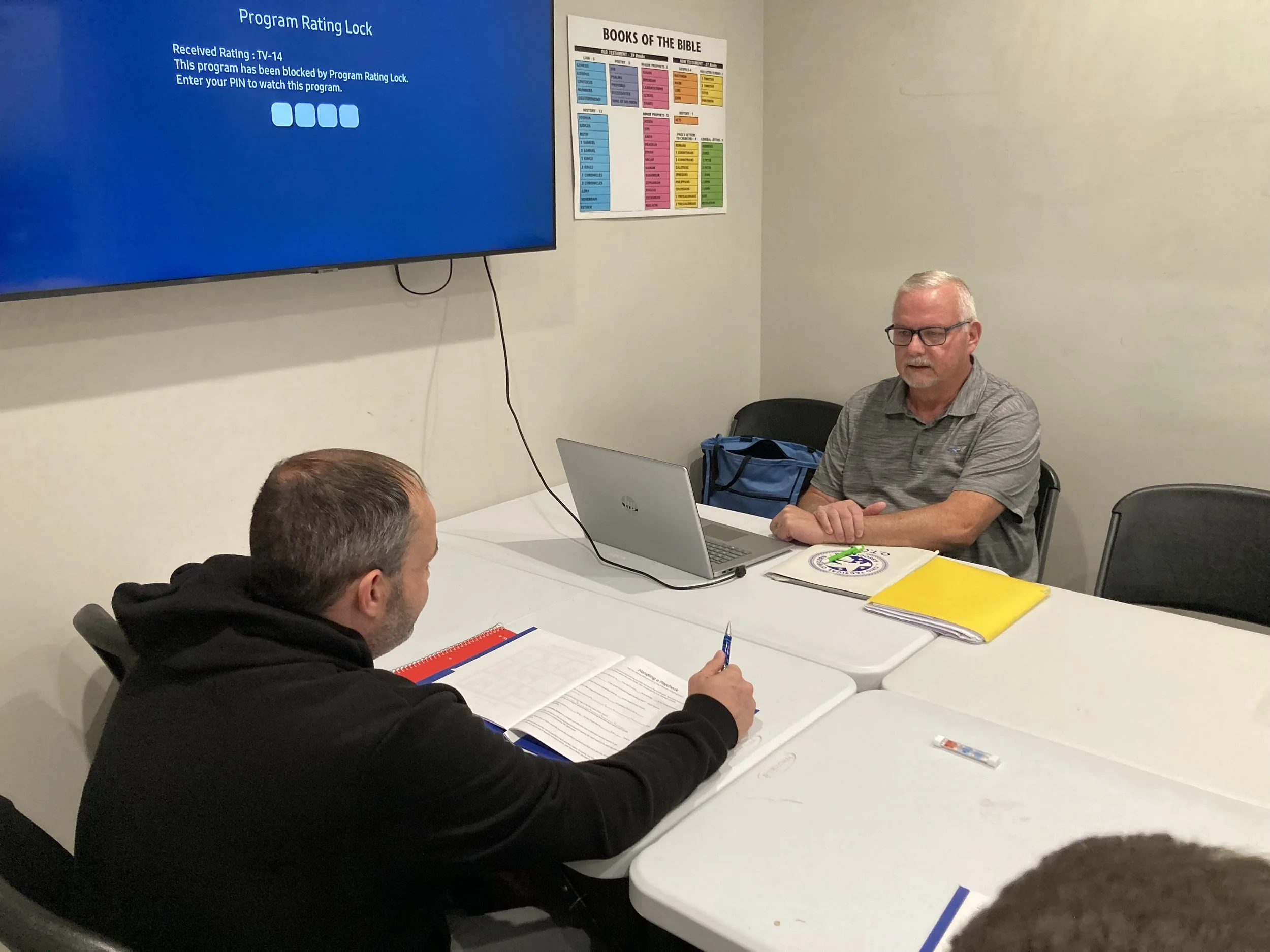 Group of people sitting around a white table in a room with a large monitor on the wall displaying a message about a program rating lock. One person is working on a laptop, and another has a notepad and pen, with medical or organizational books on the table. The room has a poster about books of the Bible on the wall.