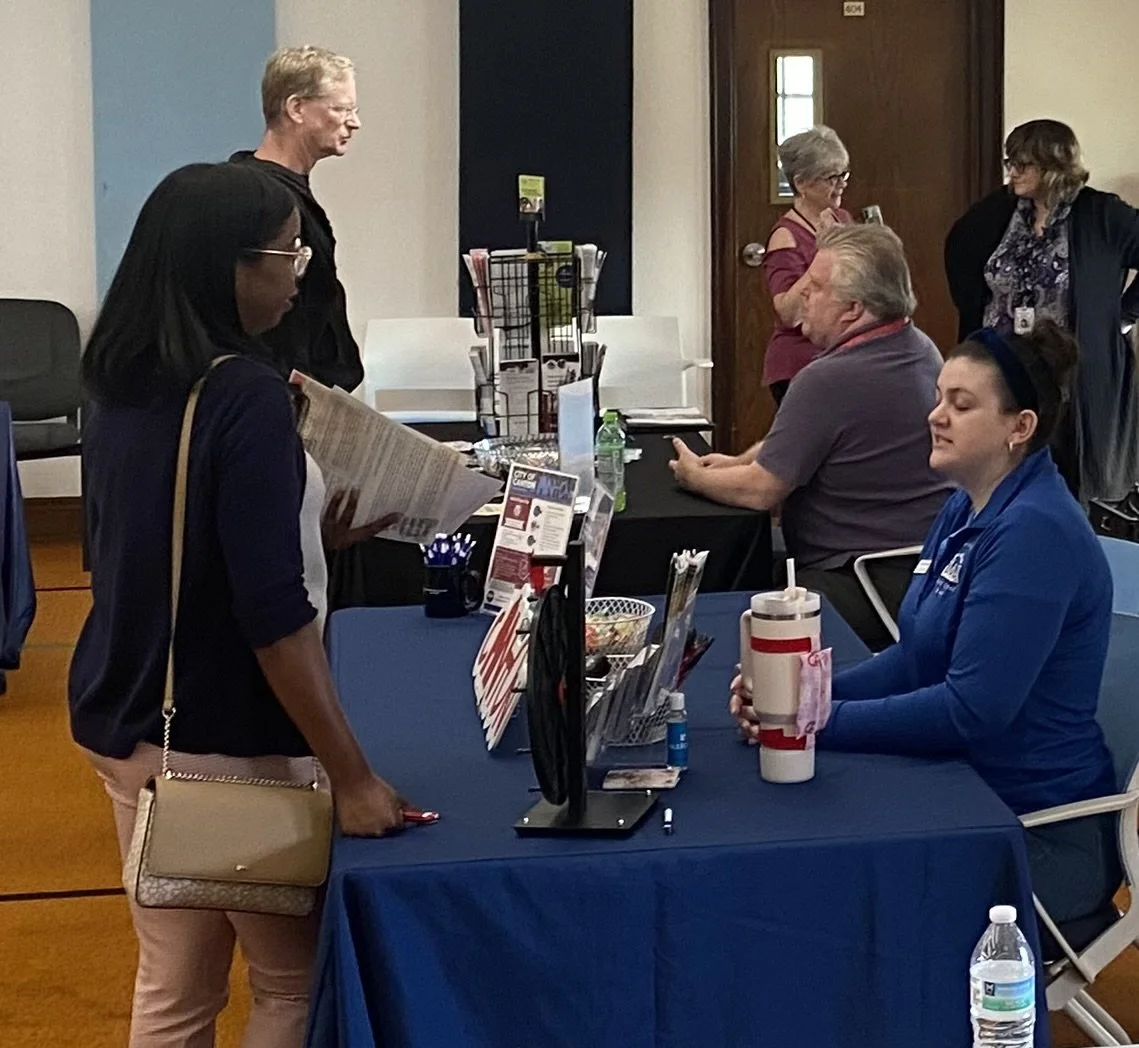 People sitting and standing at a table with informational pamphlets and bottles in a waiting or registration area.