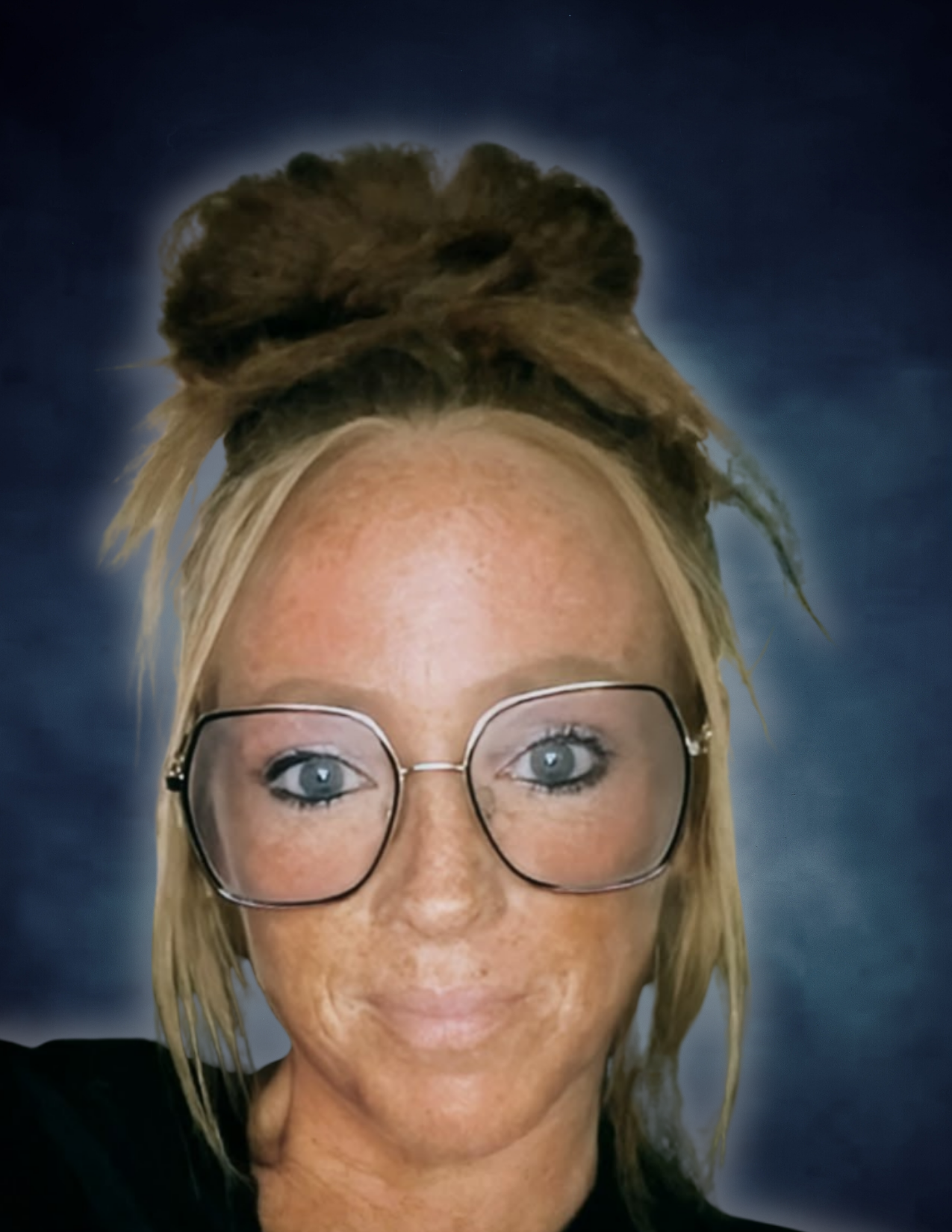 Photo of a woman with blond hair, glasses, and blue eyes, with a large brown hair bun on her head, against a dark background.