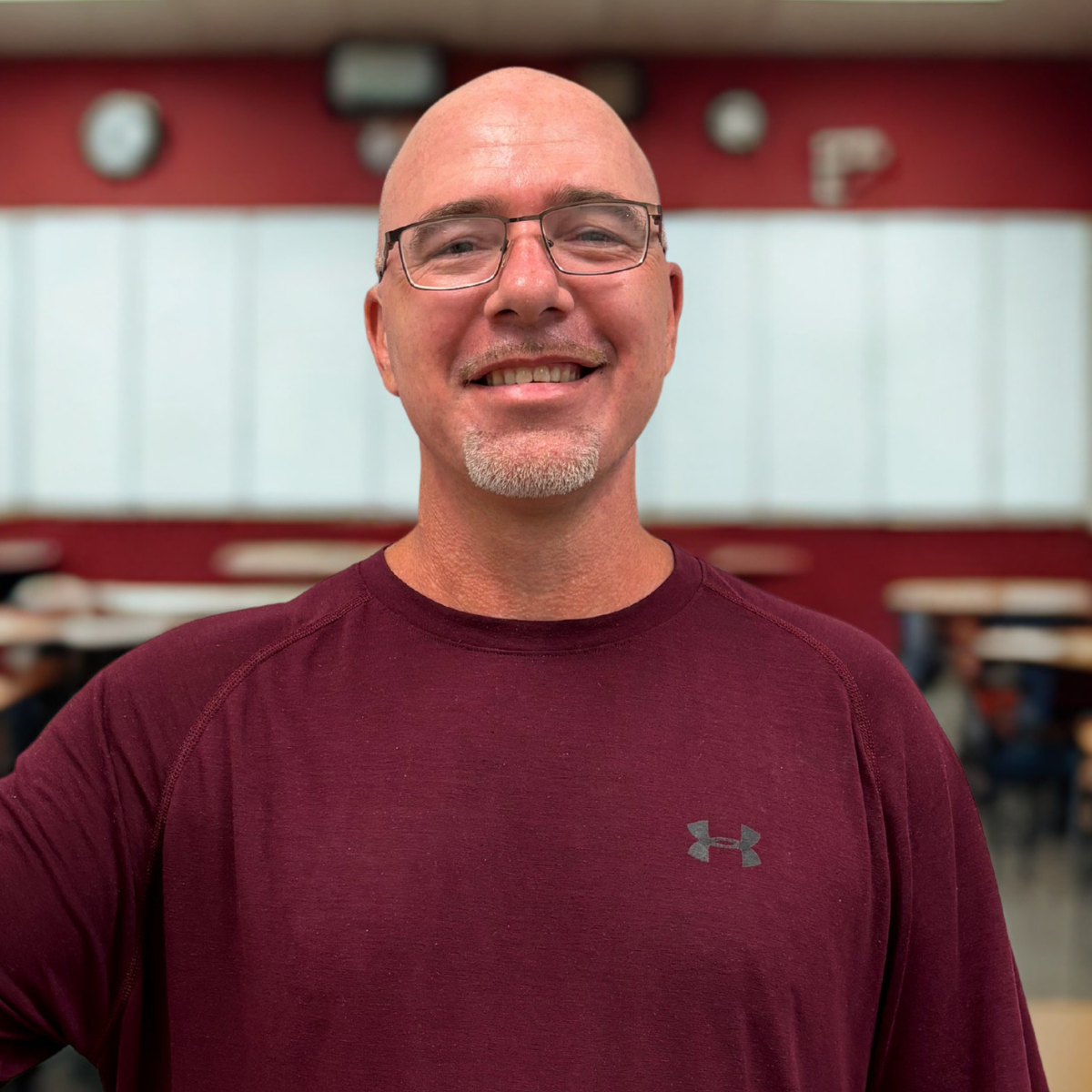 A smiling man with glasses and a goatee, wearing a maroon Under Armour long sleeve shirt, standing in a classroom or cafeteria with whiteboards and red walls in the background.
