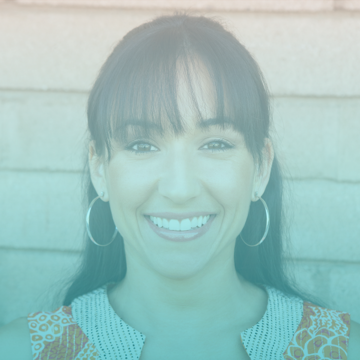 Close-up of a smiling woman with dark hair, hoop earrings, and patterned clothing, standing against a wooden wall.