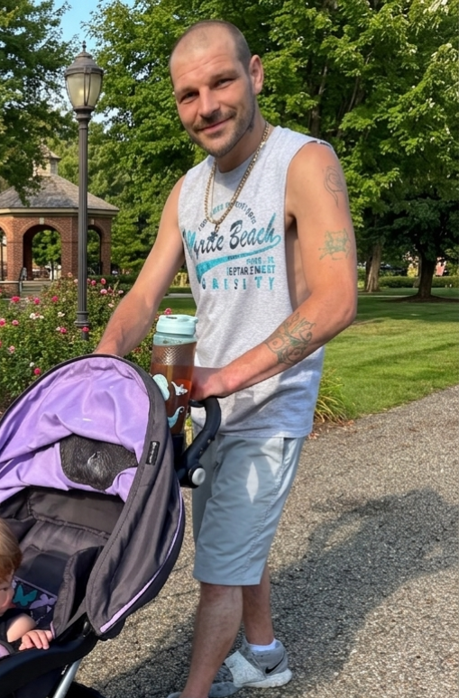 A man with a shaved head and tattoos, wearing a sleeveless gray shirt and shorts, is outdoors pushing a purple stroller with a child inside. He is smiling and holding a drink bottle. The background features green trees, a lamppost, and a brick building.