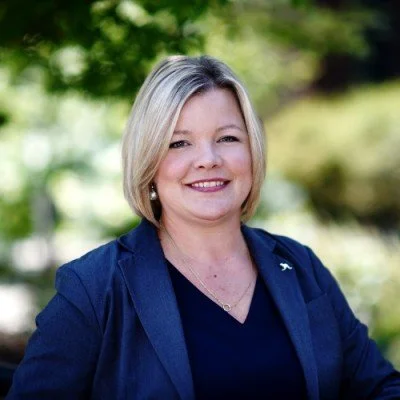 A woman with blonde hair smiling outdoors, wearing a navy blazer and top, with greenery in the background.