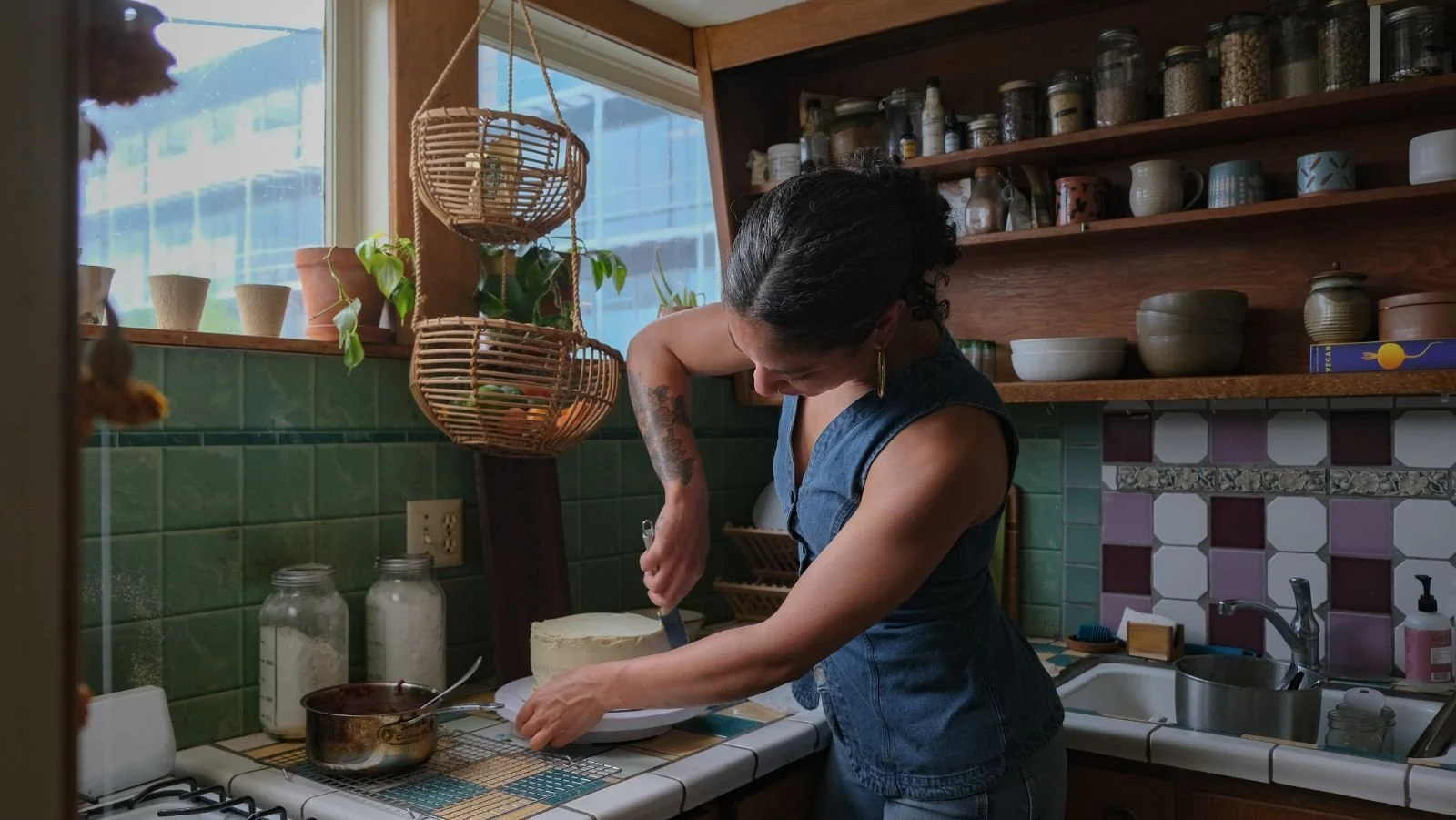 A woman with dark curly hair and tattoos on her arms is slicing a cake in a cozy kitchen with green tiled walls and wooden shelves filled with jars and cups. She is wearing a sleeveless denim top.