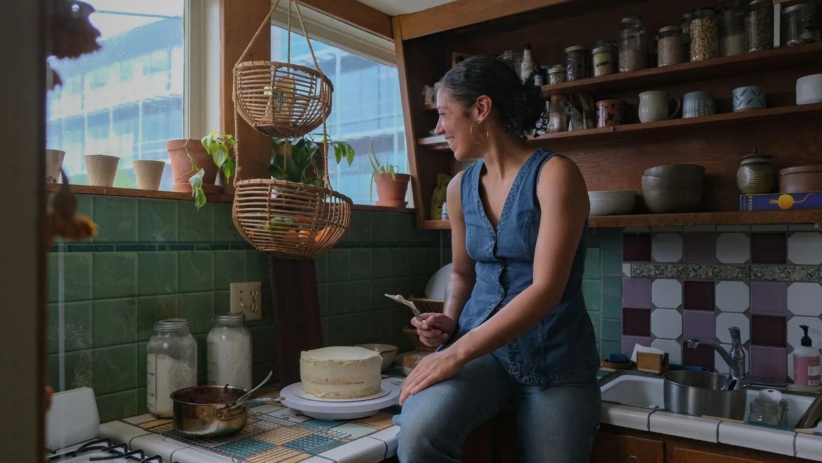 A woman sitting on a kitchen counter, cutting a cake with at least two layers in front of her. She is smiling and has her right hand resting on the counter, with her left hand holding a knife and a small piece of cake. The kitchen has green tiles, open wooden shelves with jars and cups, a window with plants, and various kitchen items.
