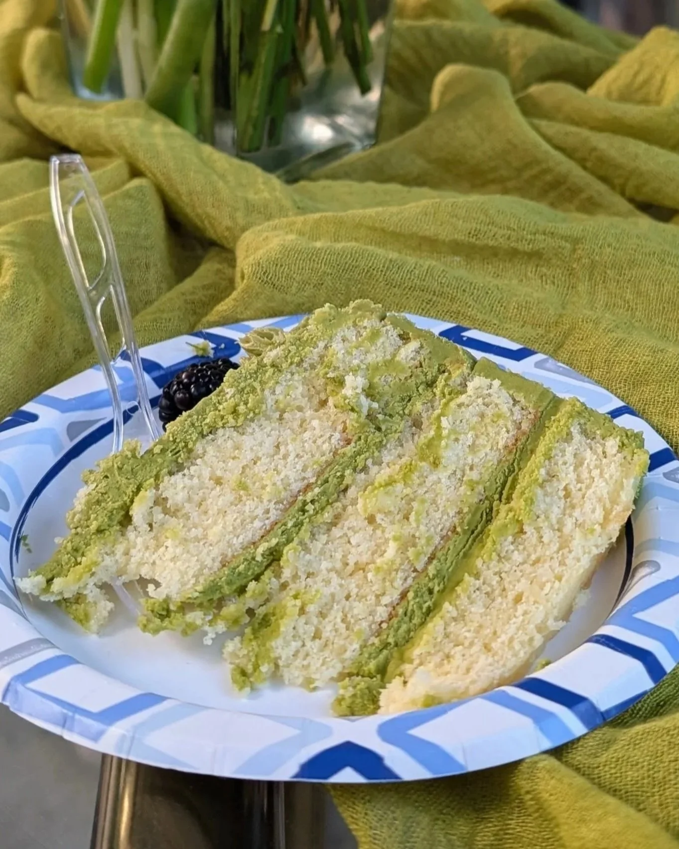 Slices of green and white layered cake with shredded white topping, served on a blue and white striped disposable plate, garnished with a blackberry, with a green cloth and a glass with greenery in the background.