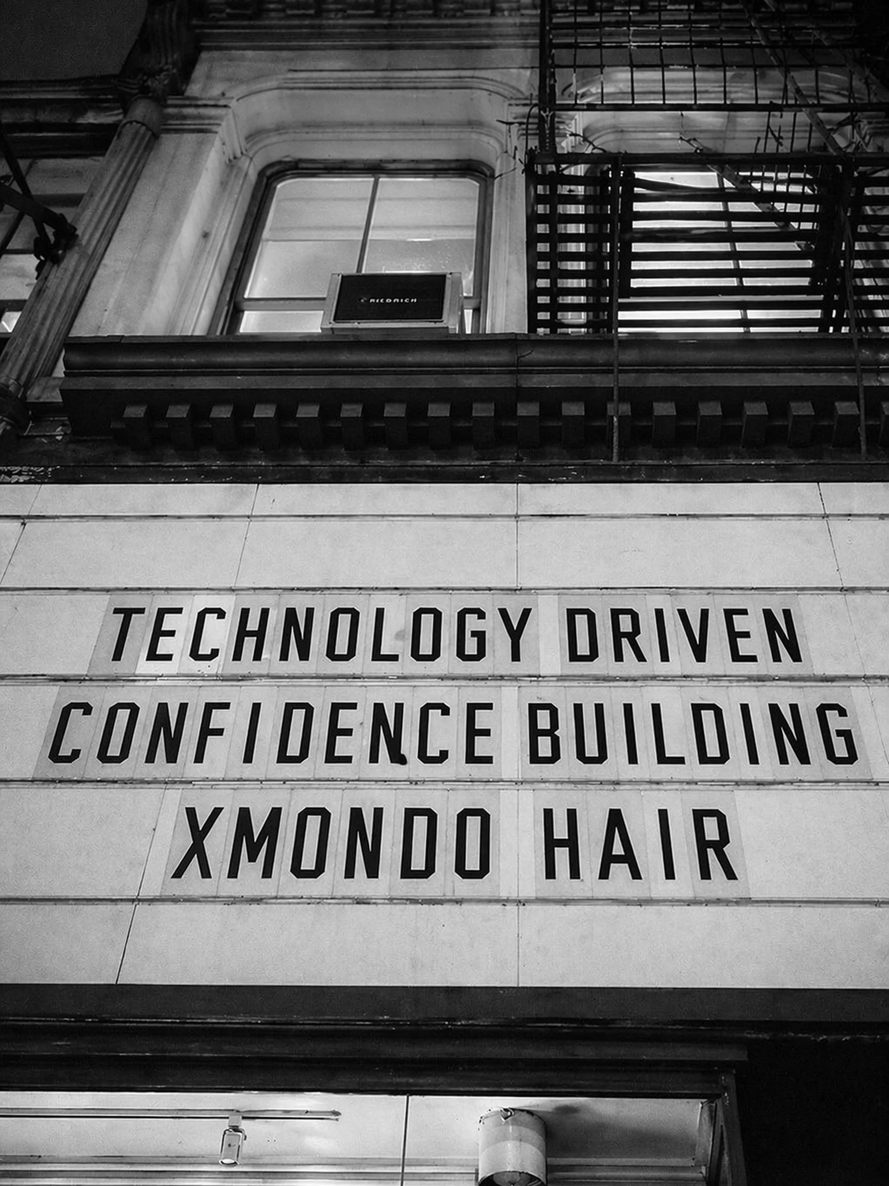 A building facade with a sign that reads "Technology Driven Confidence Building" in large letters, a window with an air conditioning unit, metal fire escape stairs, and a light fixture on the ceiling.