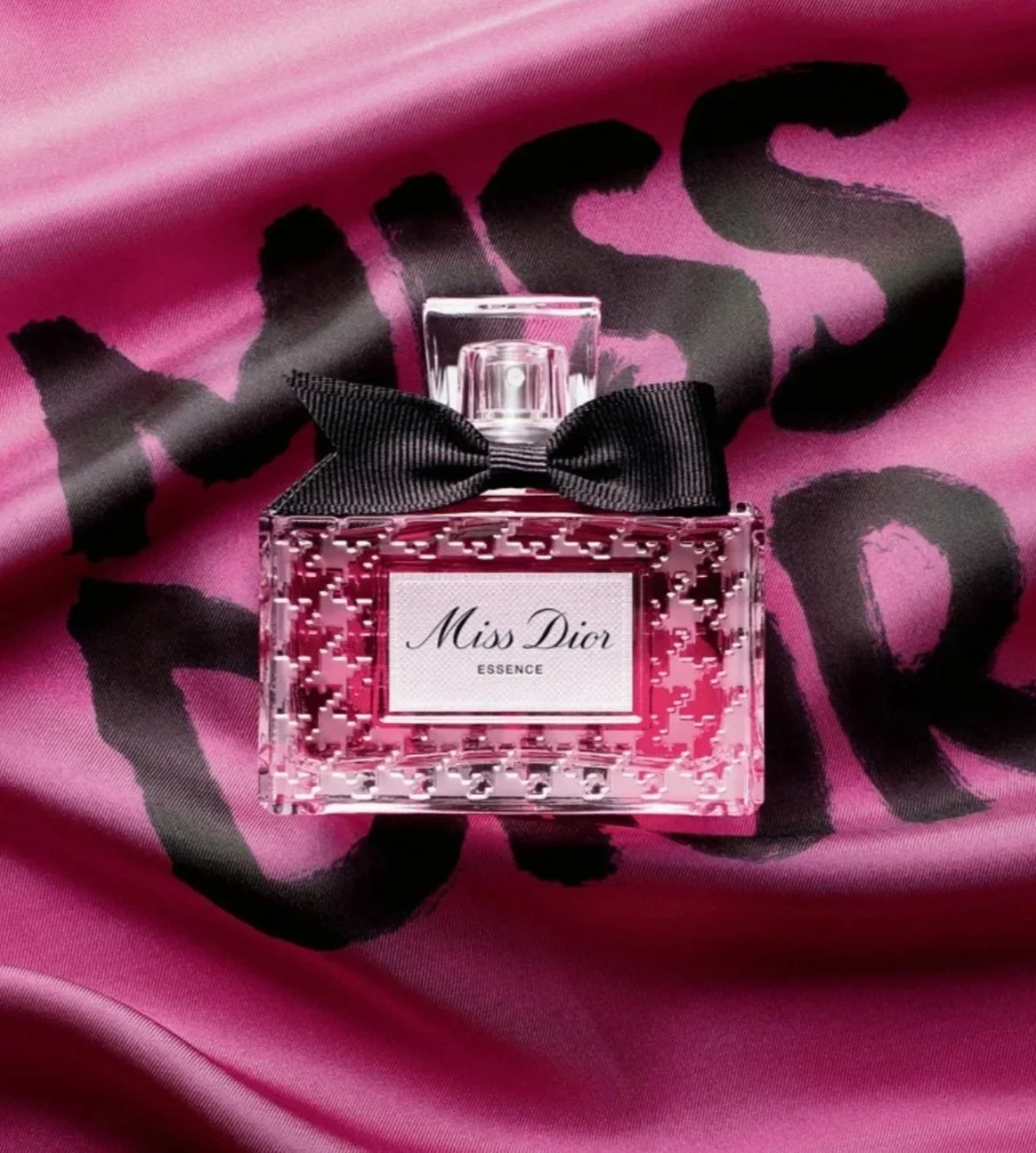 Pink perfume bottle labeled 'Miss Dior' with a black bow on top, placed on pink satin fabric with black text.