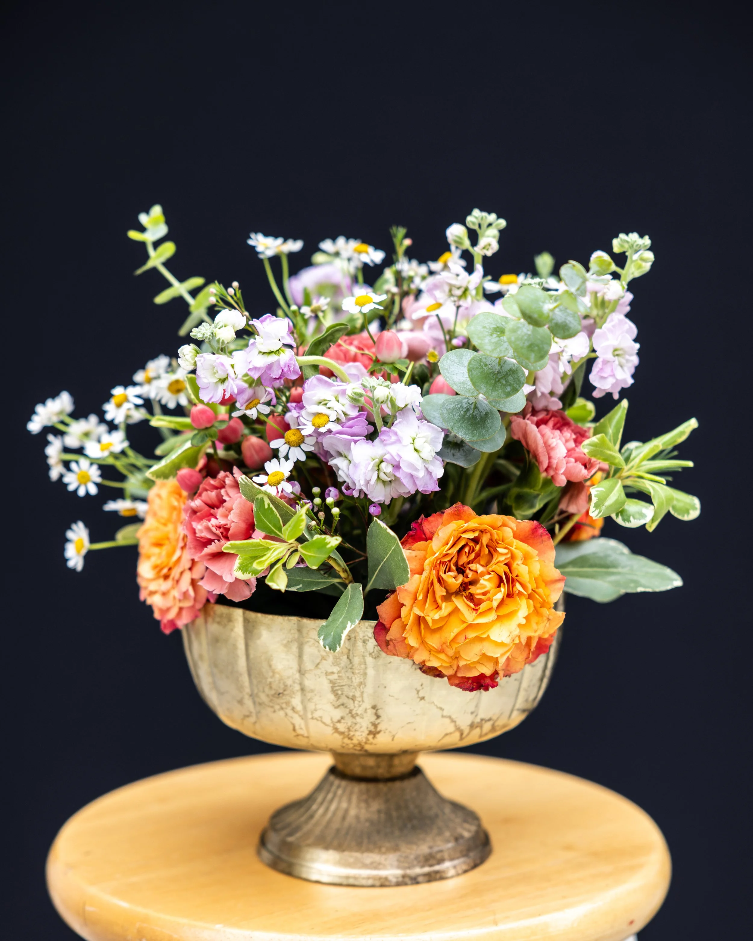 A ceramic vase with a bouquet of colorful flowers, including small daisies, pink and orange roses, and green foliage, set on a round wooden stand against a black background.