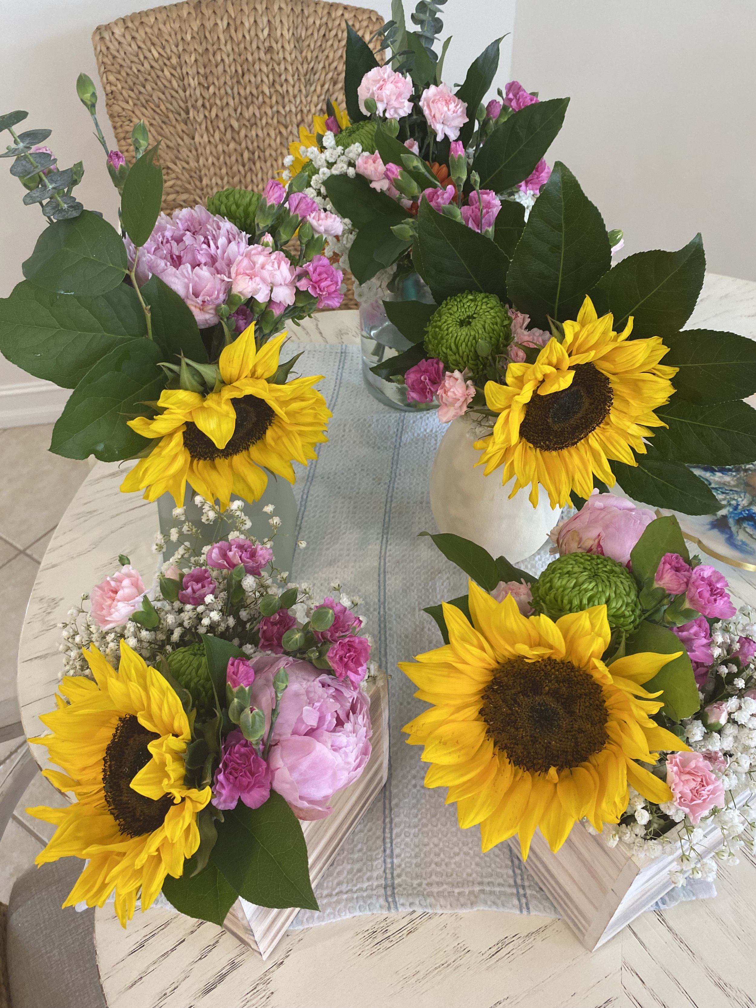 Two floral arrangements featuring sunflowers, pink carnations, and small white flowers on a light-colored table.