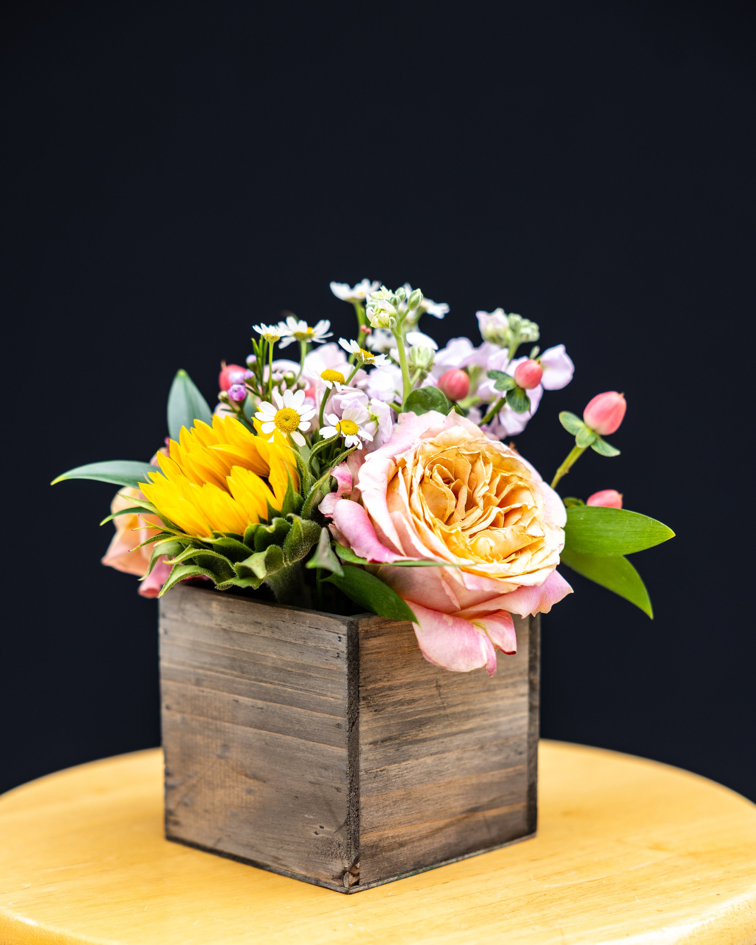 A bouquet of mixed flowers, including sunflowers, roses, and daisies, in a rustic wooden box on a yellow table with a black background.