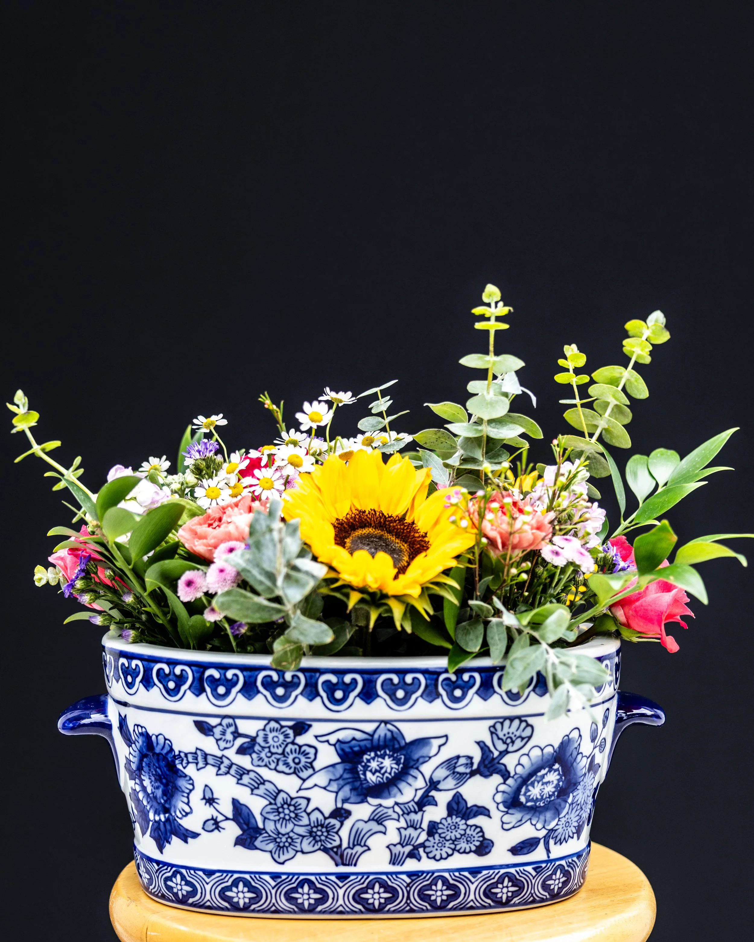 A colorful bouquet of various flowers, including a large sunflower, in a blue and white patterned ceramic vase on a wooden surface against a black background.