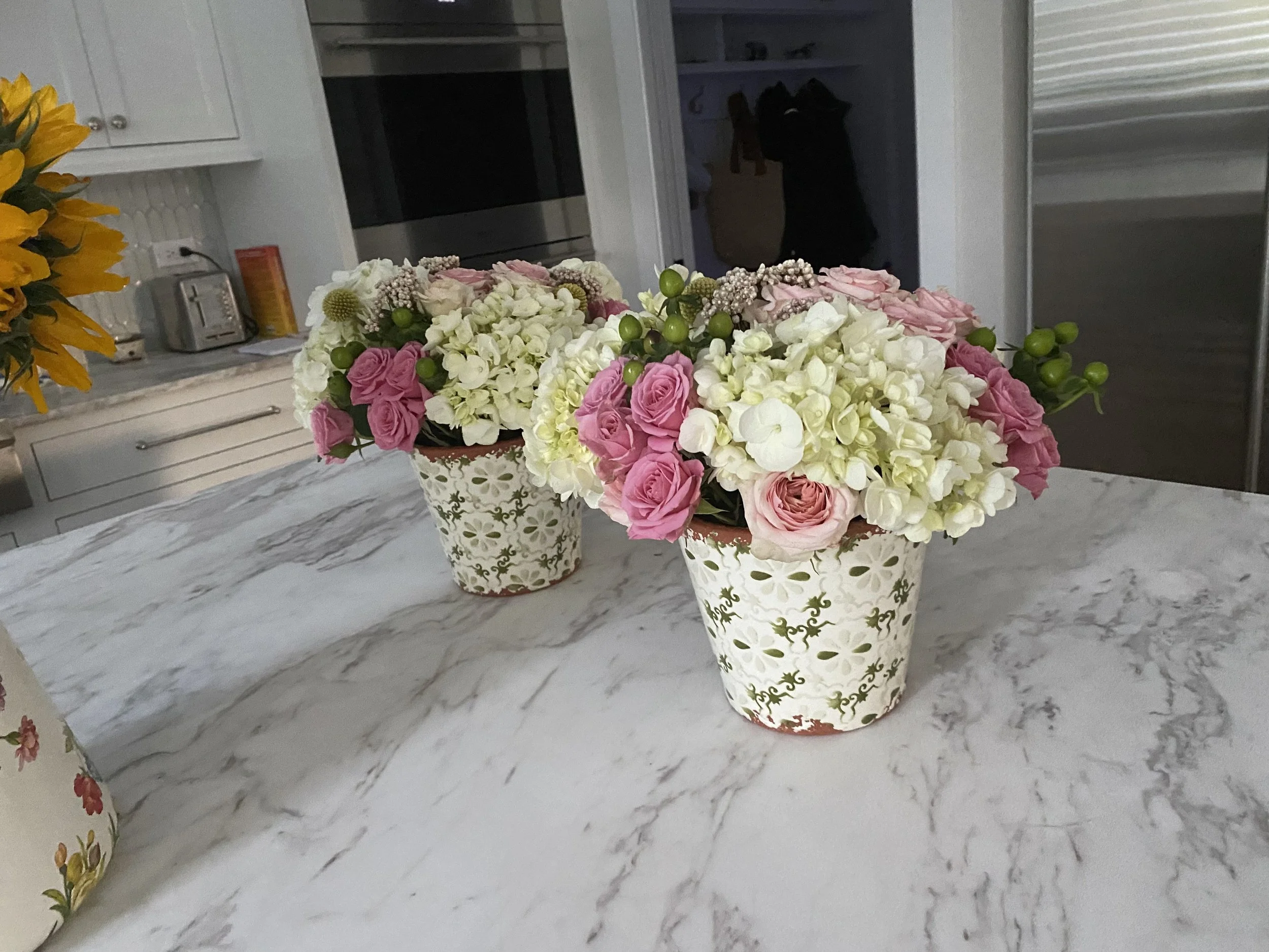 Three decorative flower pots filled with pink and white roses, hydrangeas, and greenery on a marble countertop in a kitchen.