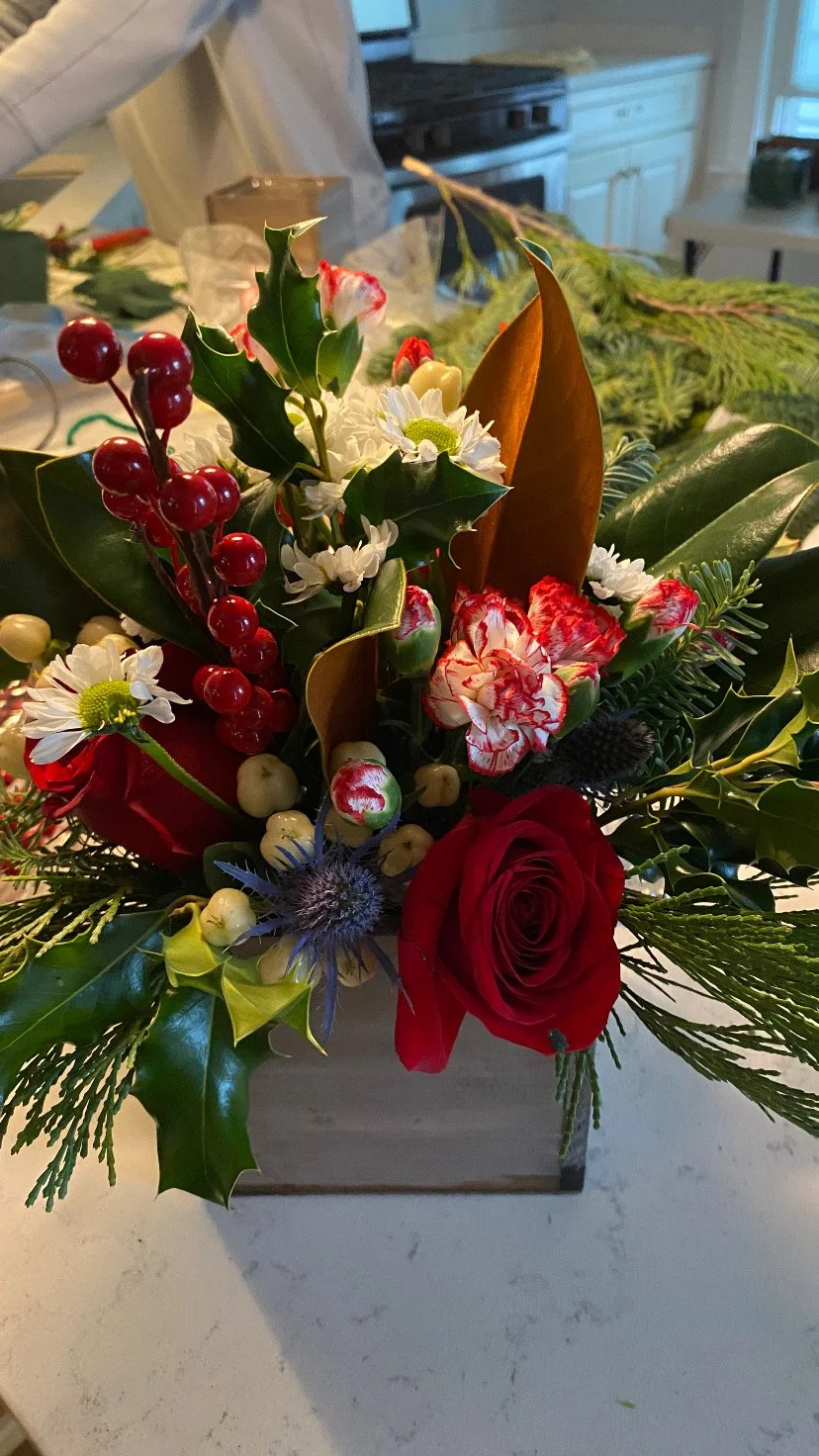 A festive floral arrangement with red roses, holly leaves, white daisies, red berries, and other greenery, in a wooden container, on a kitchen countertop.