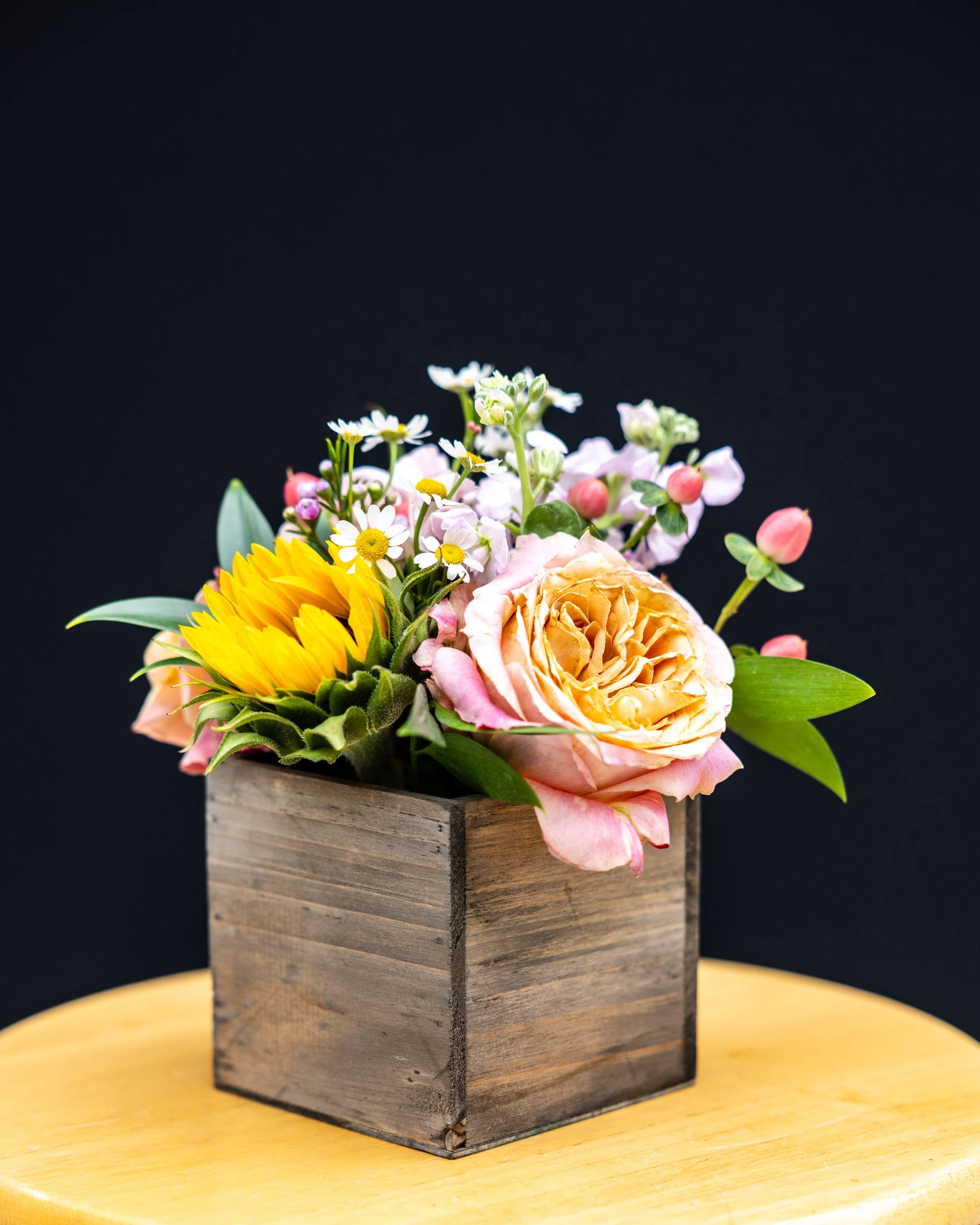 A bouquet of mixed flowers including sunflowers, roses, and daisies arranged in a wooden box on a yellow table against a black background.