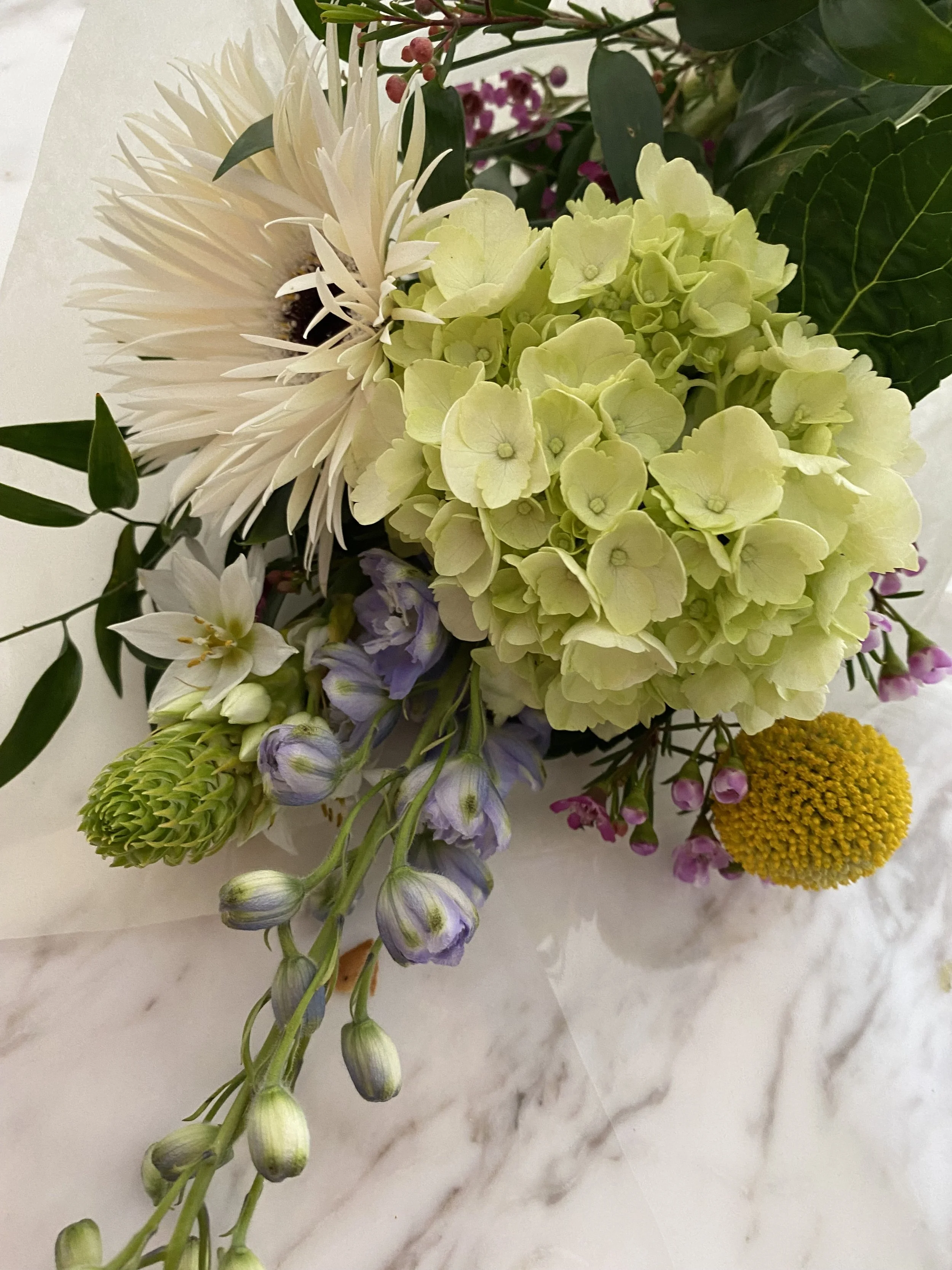 Close-up of a floral arrangement with white, green, purple, and yellow flowers on a white marble surface.