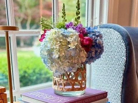 A bouquet of colorful flowers in a glass vase, placed on a stack of books next to a window.