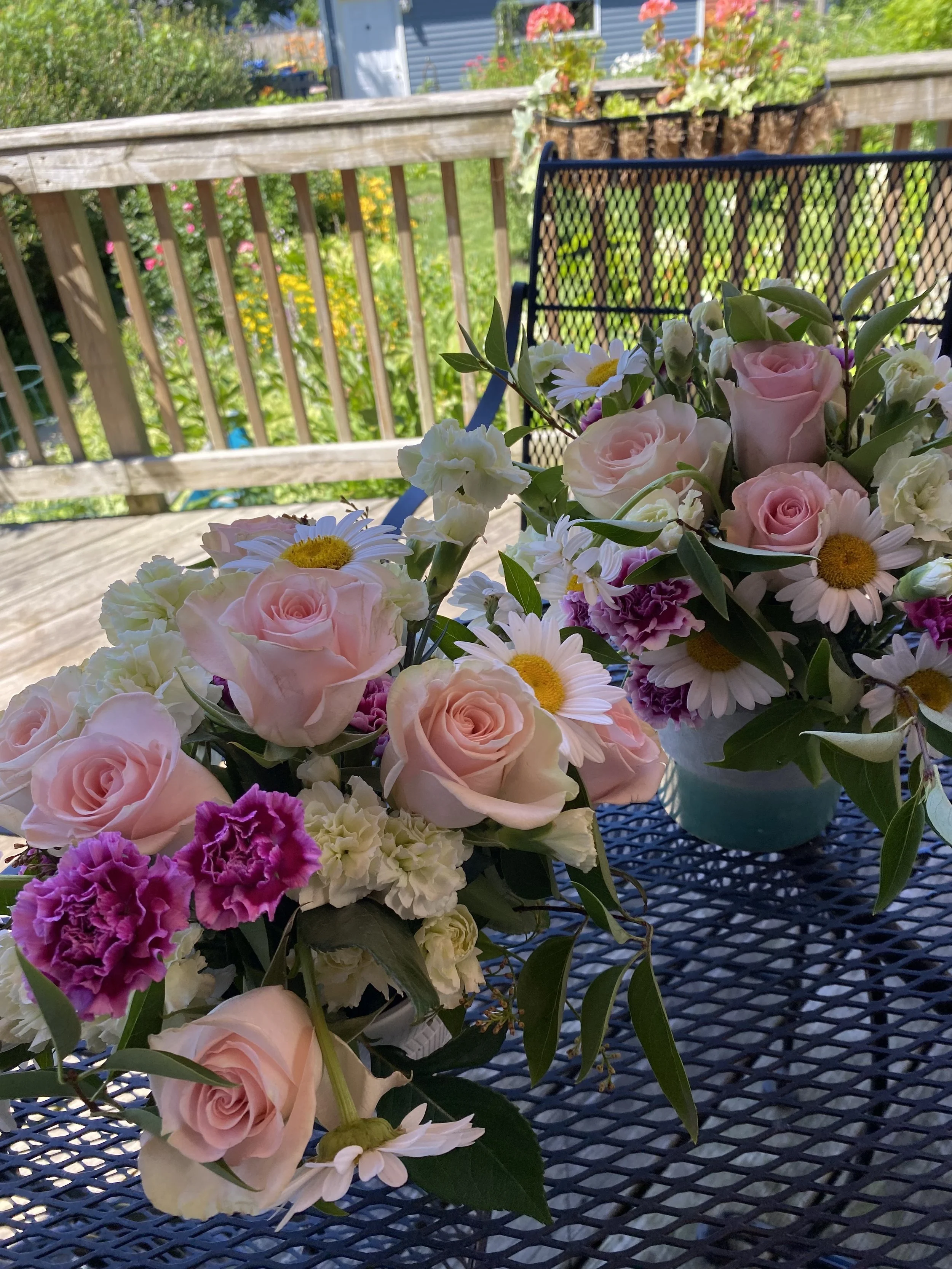 Close-up of a bouquet of pink and white roses, daisies, and other flowers on a blue outdoor table. In the background, a wooden railing with flower pots and a garden with colorful flowers and a blue house.