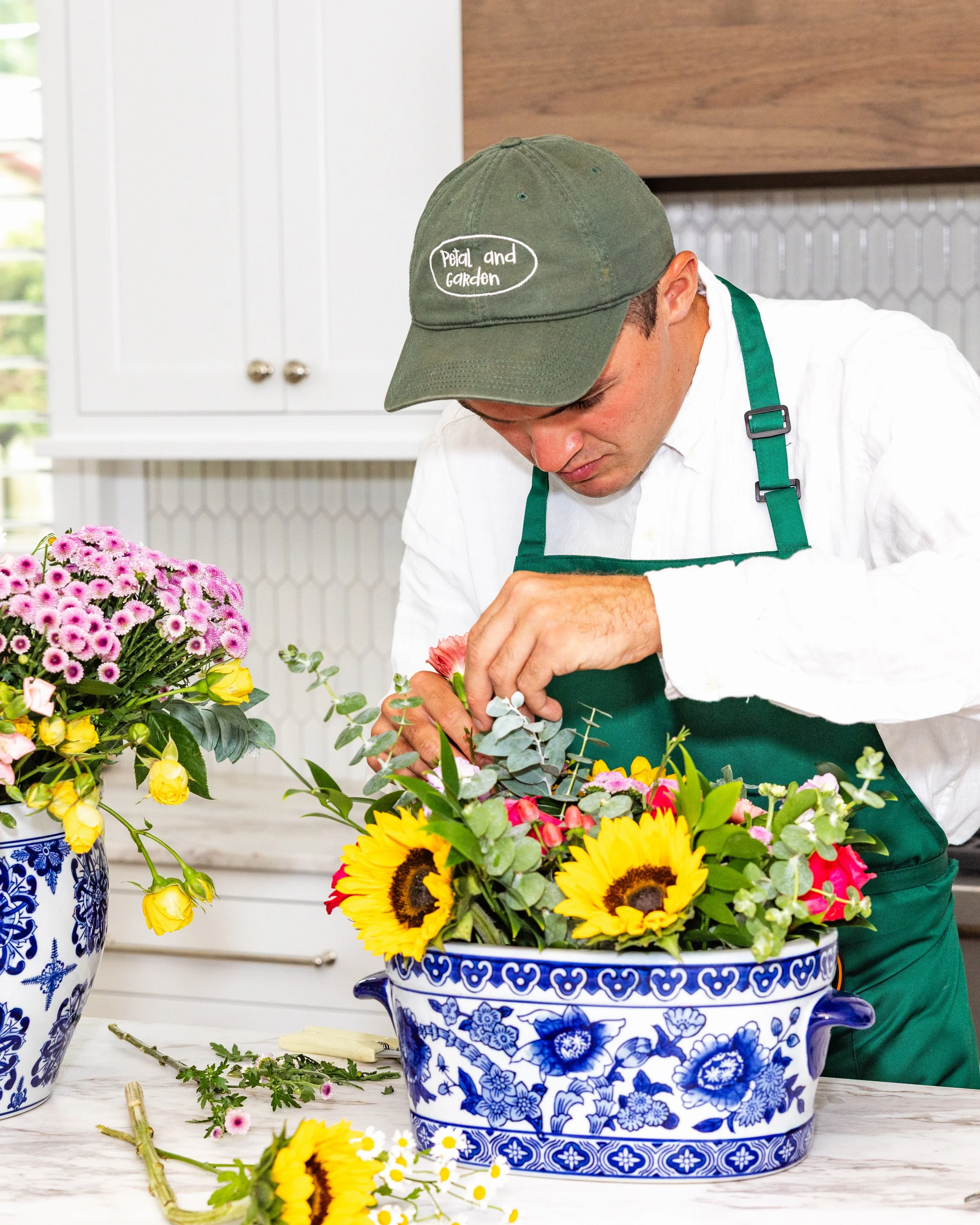 A person arranging a colorful flower bouquet in decorative blue-and-white ceramic vases. They are wearing a green cap with white writing that says 'Petal and Garden' and a green apron over white shirt, in a kitchen setting.