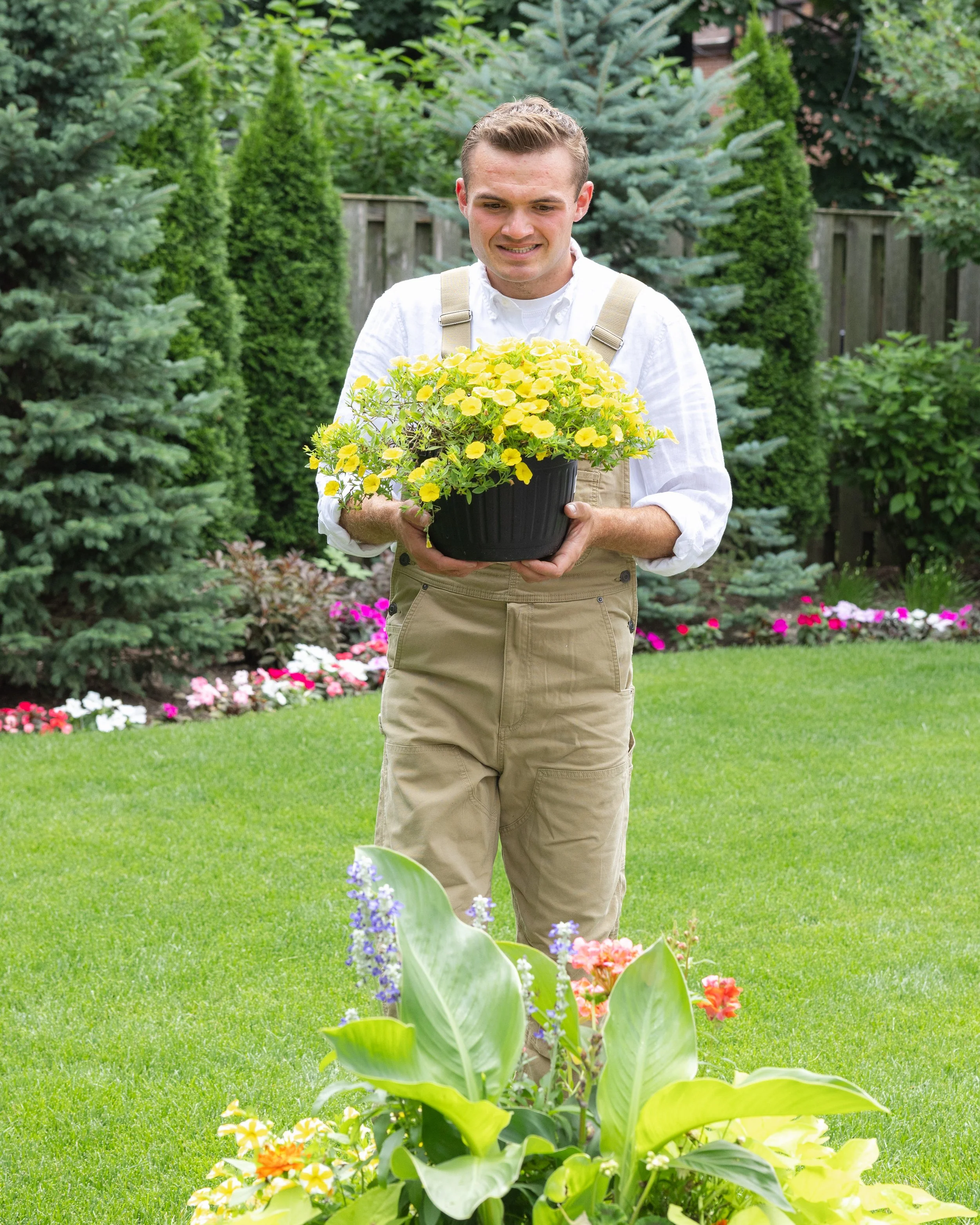 Sean holding a pot with yellow flowers in a garden surrounded by green plants and trees that he planted.