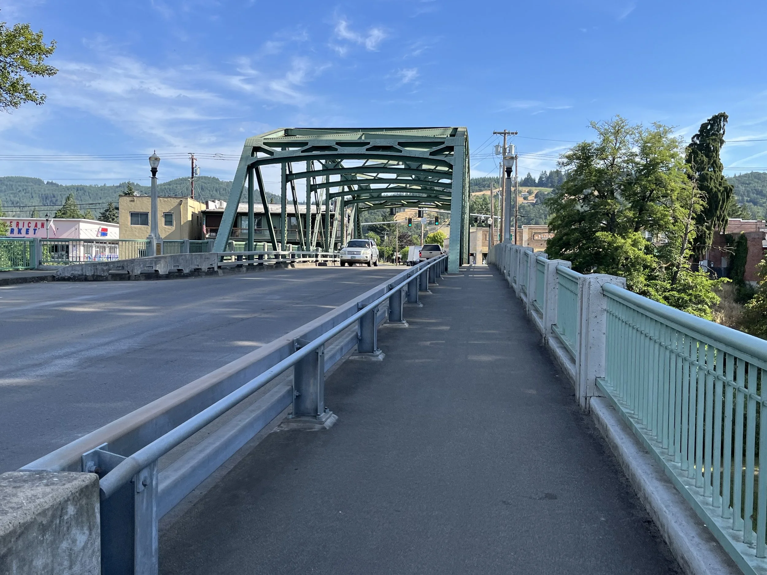 View of a metal bridge with a pedestrian walkway and railing on the right. The bridge is over a road, with buildings and trees in the background under a blue sky with some clouds.