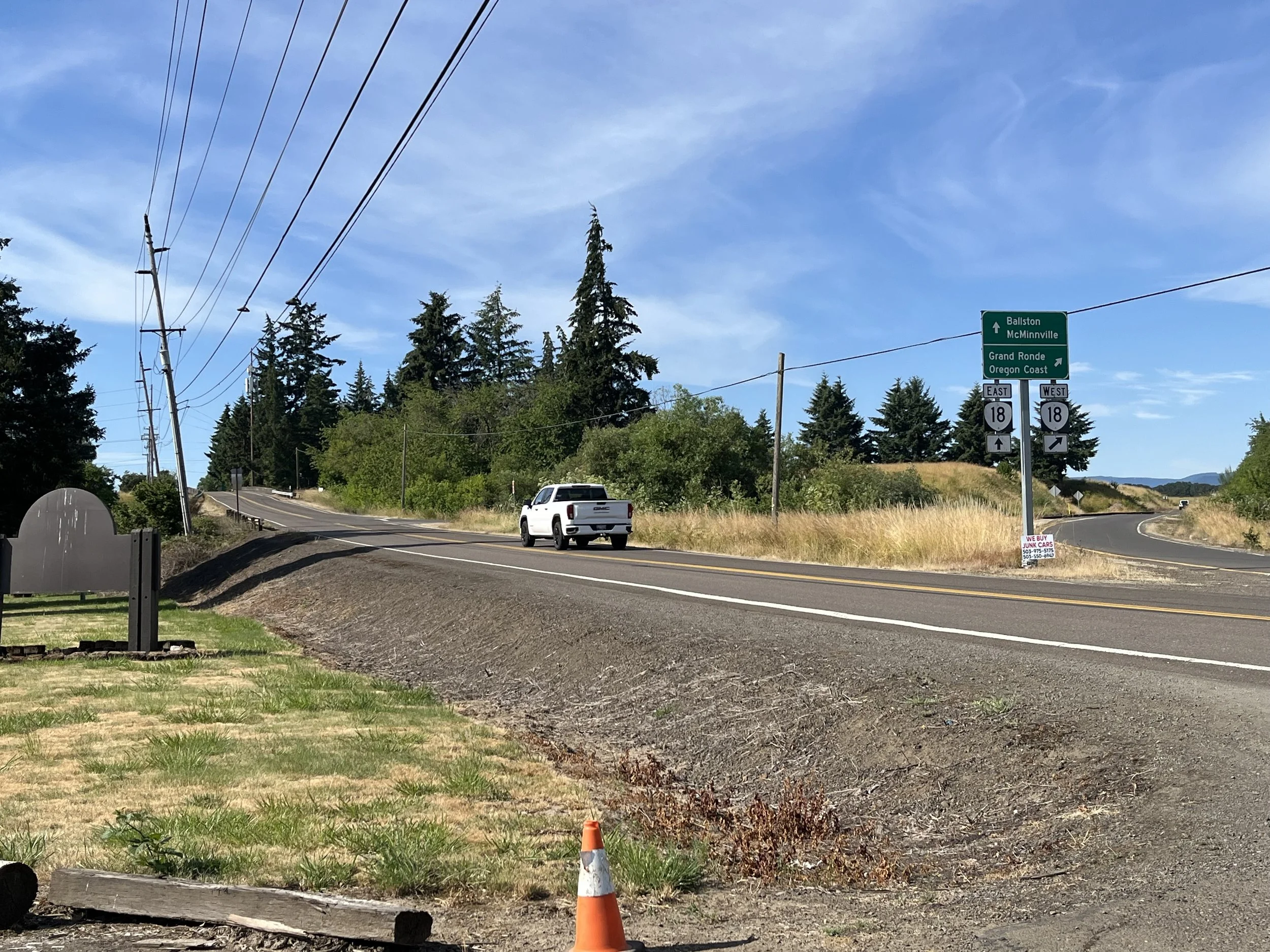 A two-lane road with a white truck driving past a green highway sign indicating directions to Ballston, McMinnville, Grand Ronde, and Oregon Coast. There are power lines on the left side of the road, tall trees, and a grassy area with a small orange traffic cone near the bottom of the image. The sky is clear with some wispy clouds.