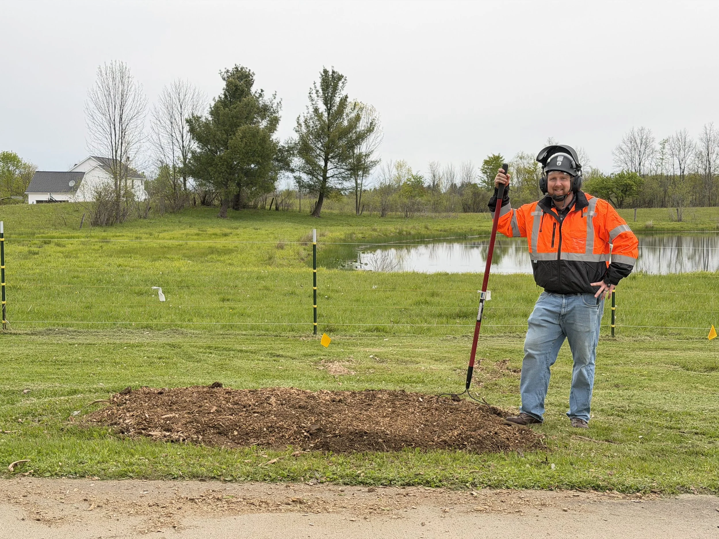 A man wearing an orange safety jacket and black helmet is holding a gardening tool near a freshly dug hole in a grassy field with trees and a house in the background.