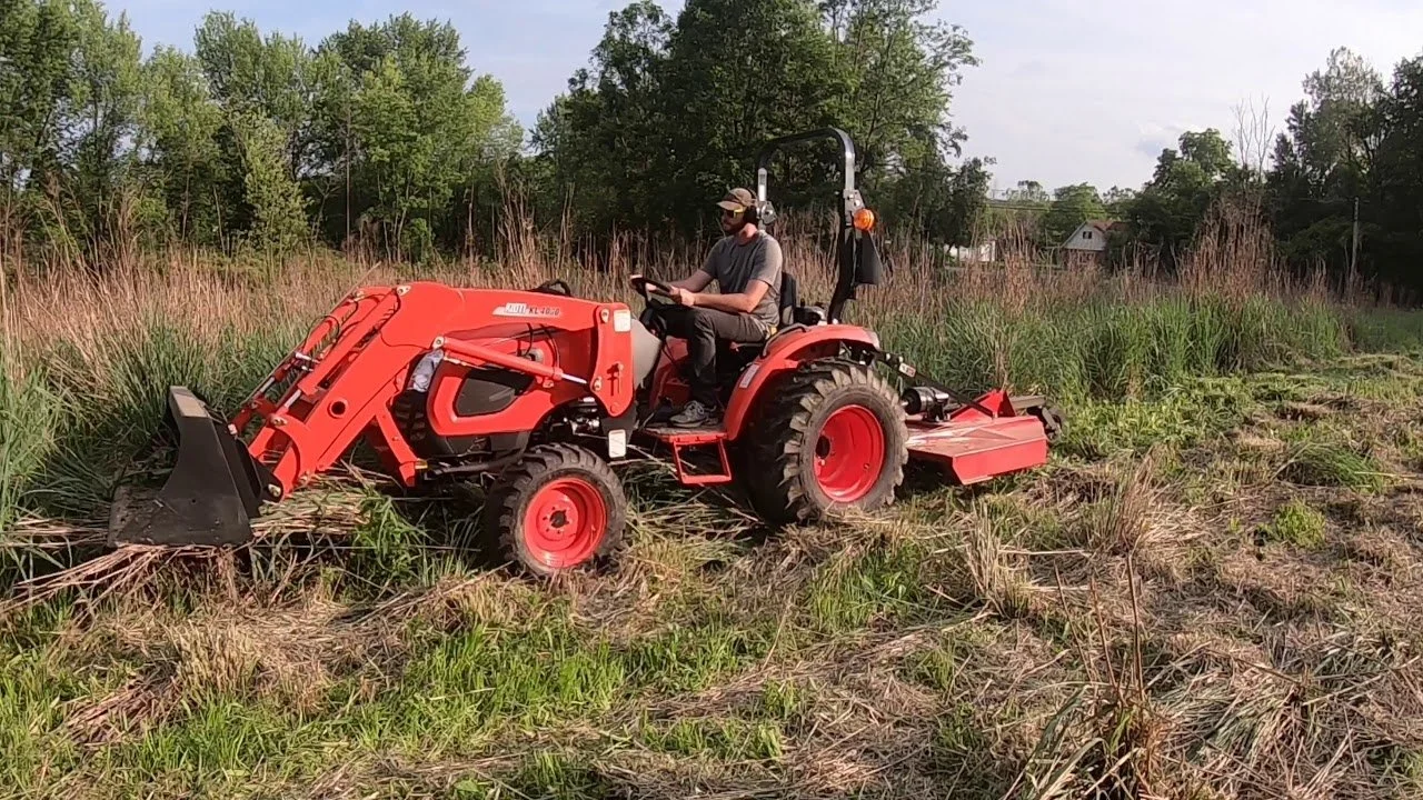 A person is driving a red tractor with a front loader attachment in a grassy, rural field surrounded by trees.