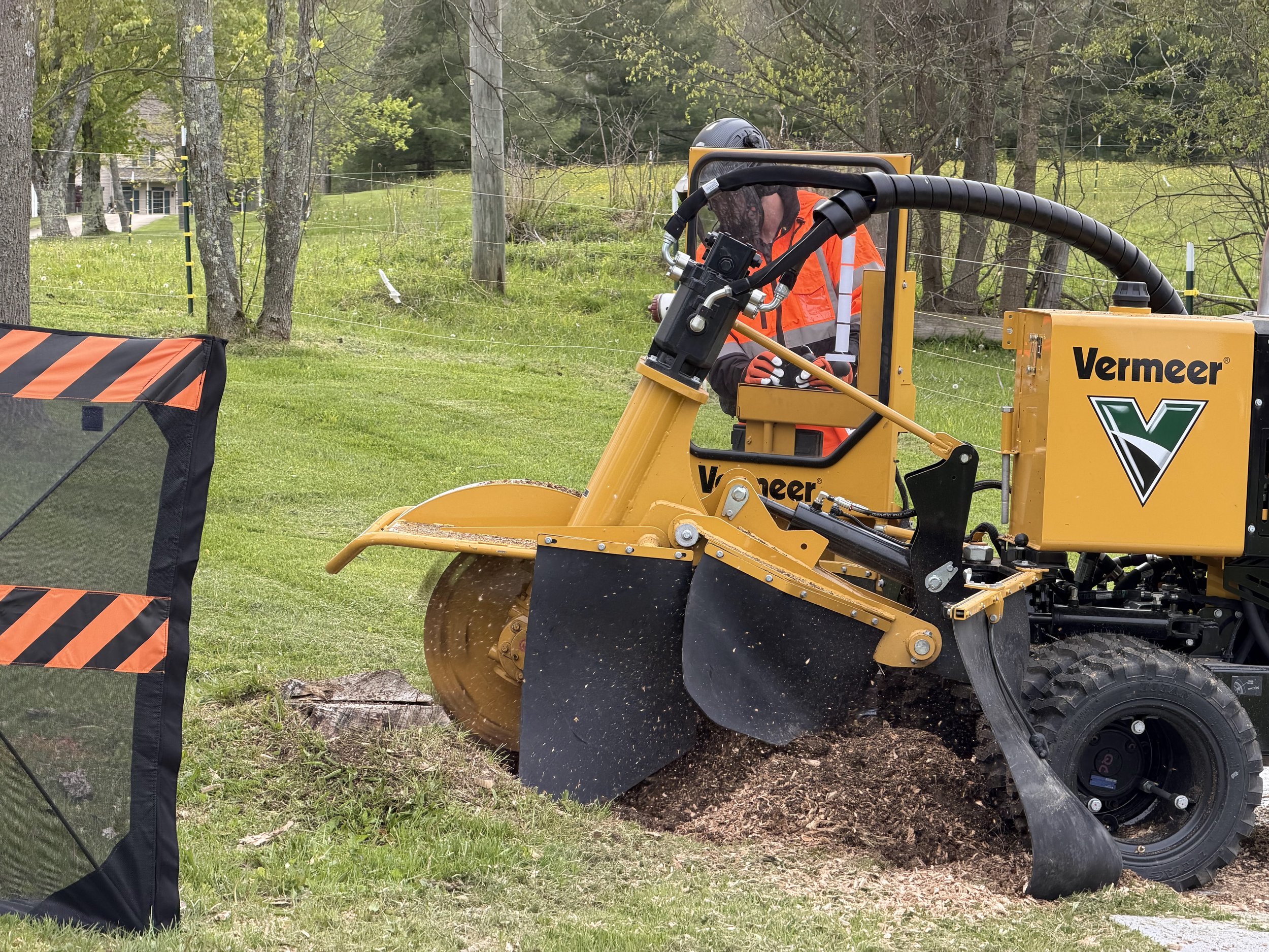 A worker operating a yellow Vermeer stump grinder, cutting a tree stump in a grassy area with fence posts and trees in the background.