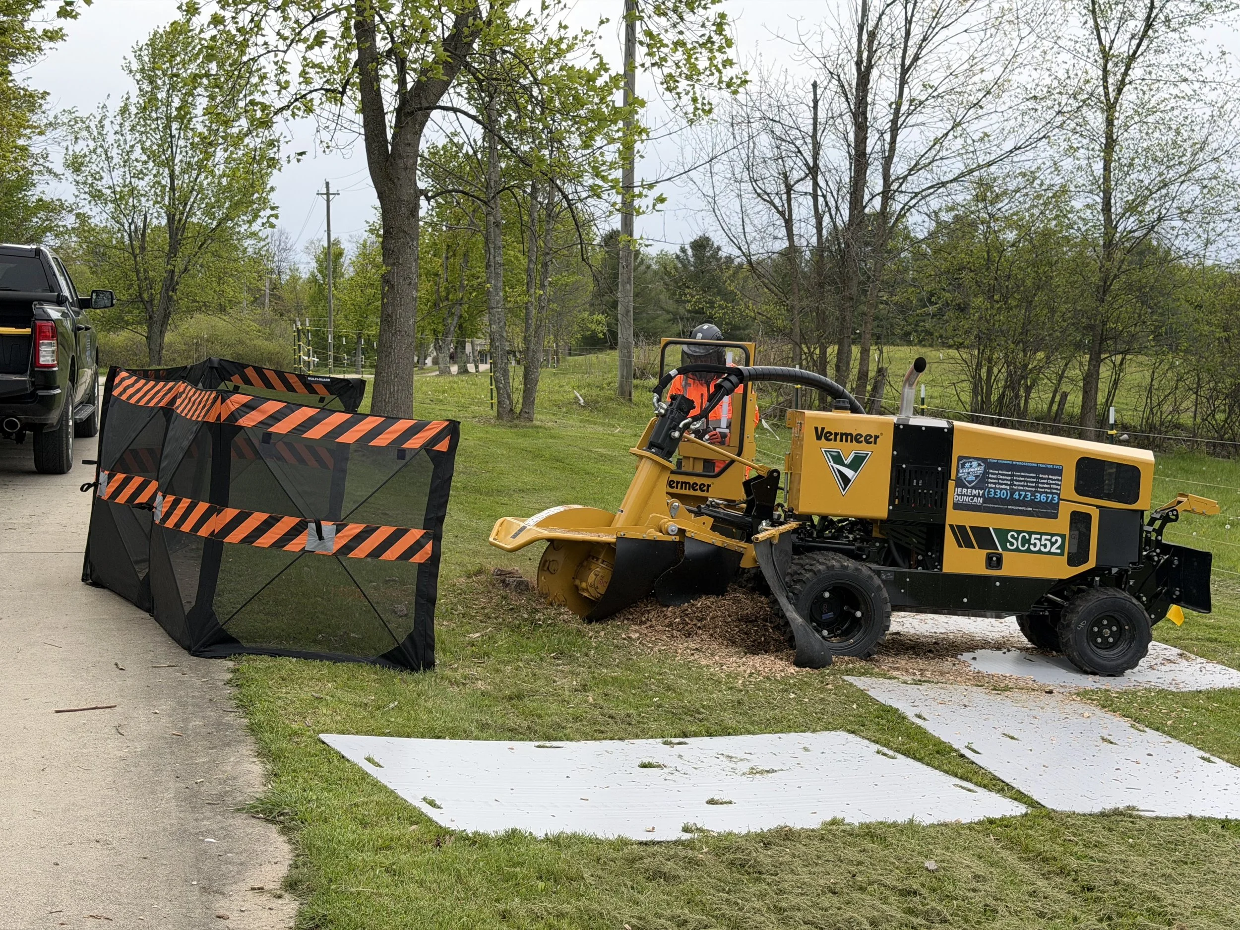 Construction worker operating a compacting machine on a sidewalk, with safety barriers and trees in the background.