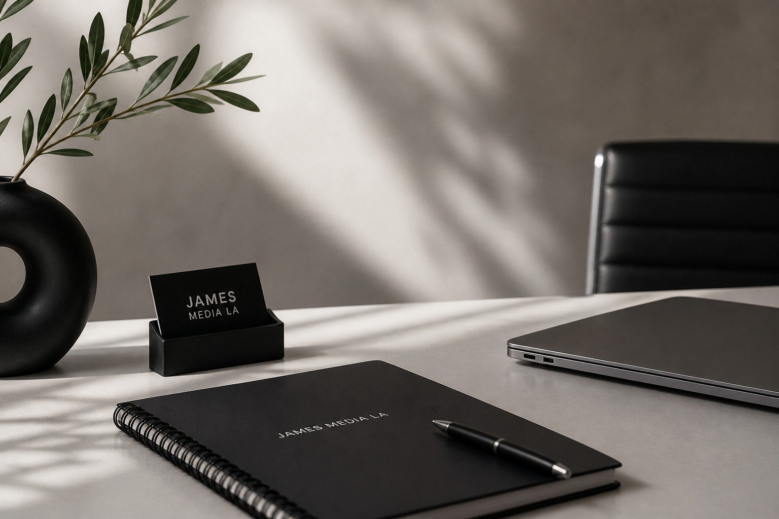 A minimalist office desk setup with a potted olive branch, a black business card holder with the name "James Media LA," a black spiral notebook with matching pen, and a closed silver laptop, all arranged on a smooth, gray surface with shadowed background.