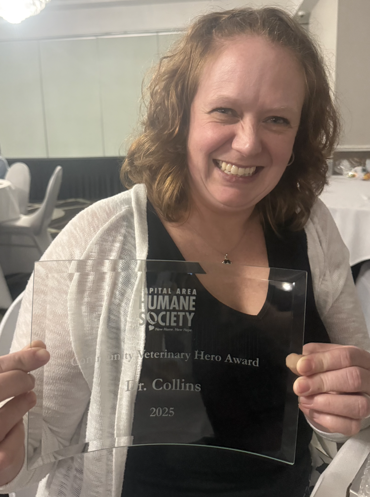 A woman with curly brown hair smiling and holding an award plaque that reads 'Community Veterinary Hero Award' from the Capital Area Humane Society, in a banquet hall.