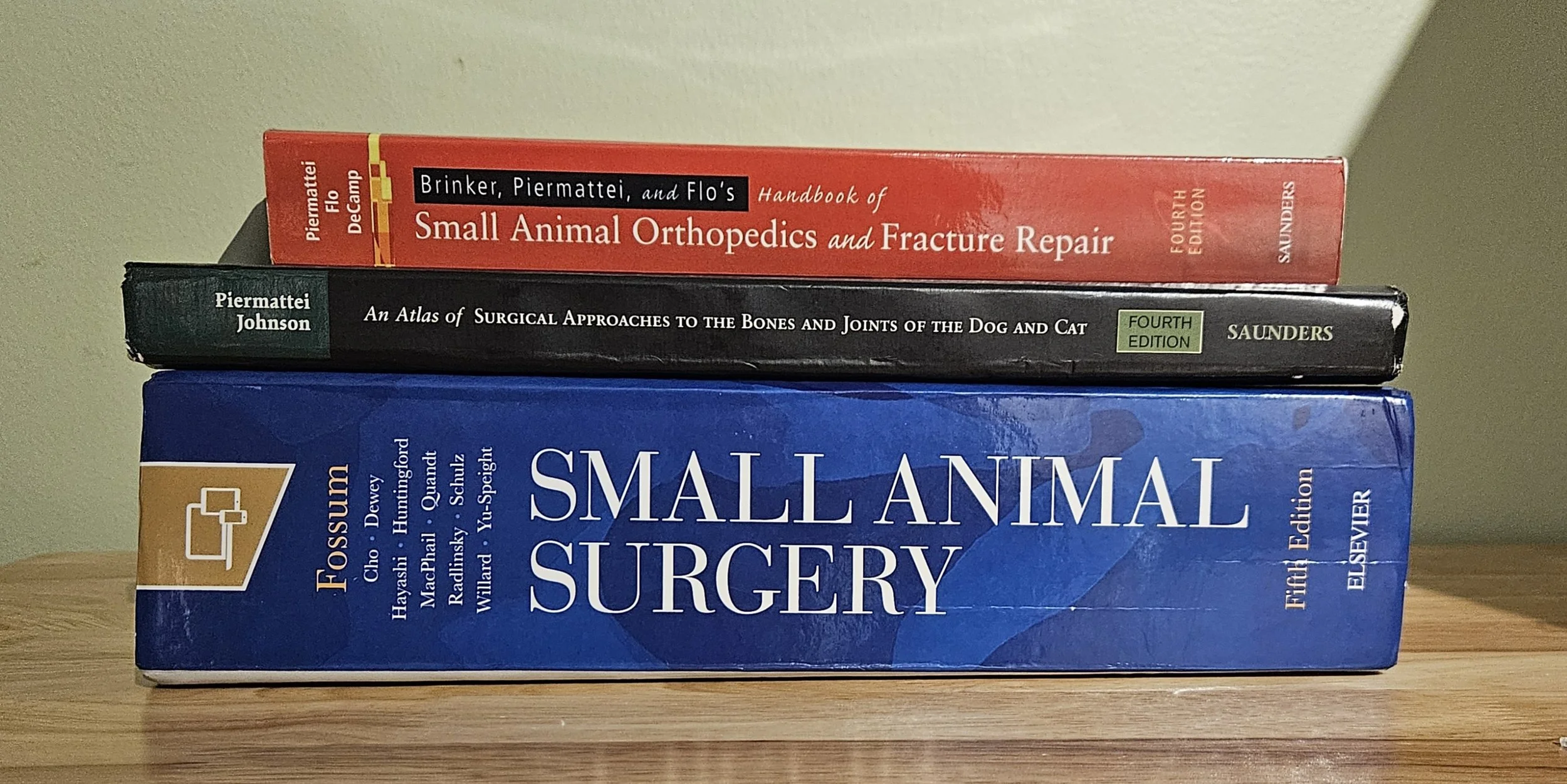 Stack of three veterinary medical books on small animal surgery, orthopedic fracture repair, and surgical approaches to bones and joints, placed on a wooden surface against a light-colored wall.