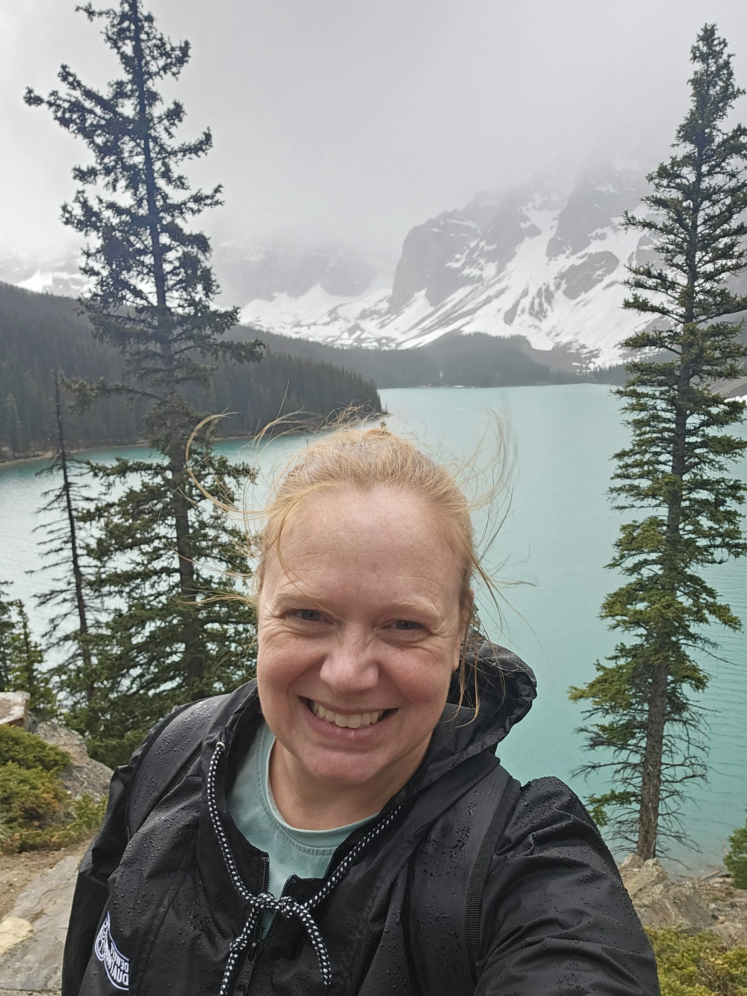 A woman smiling in a rain jacket outdoors near a lake with snow-capped mountains and evergreen trees in the background.