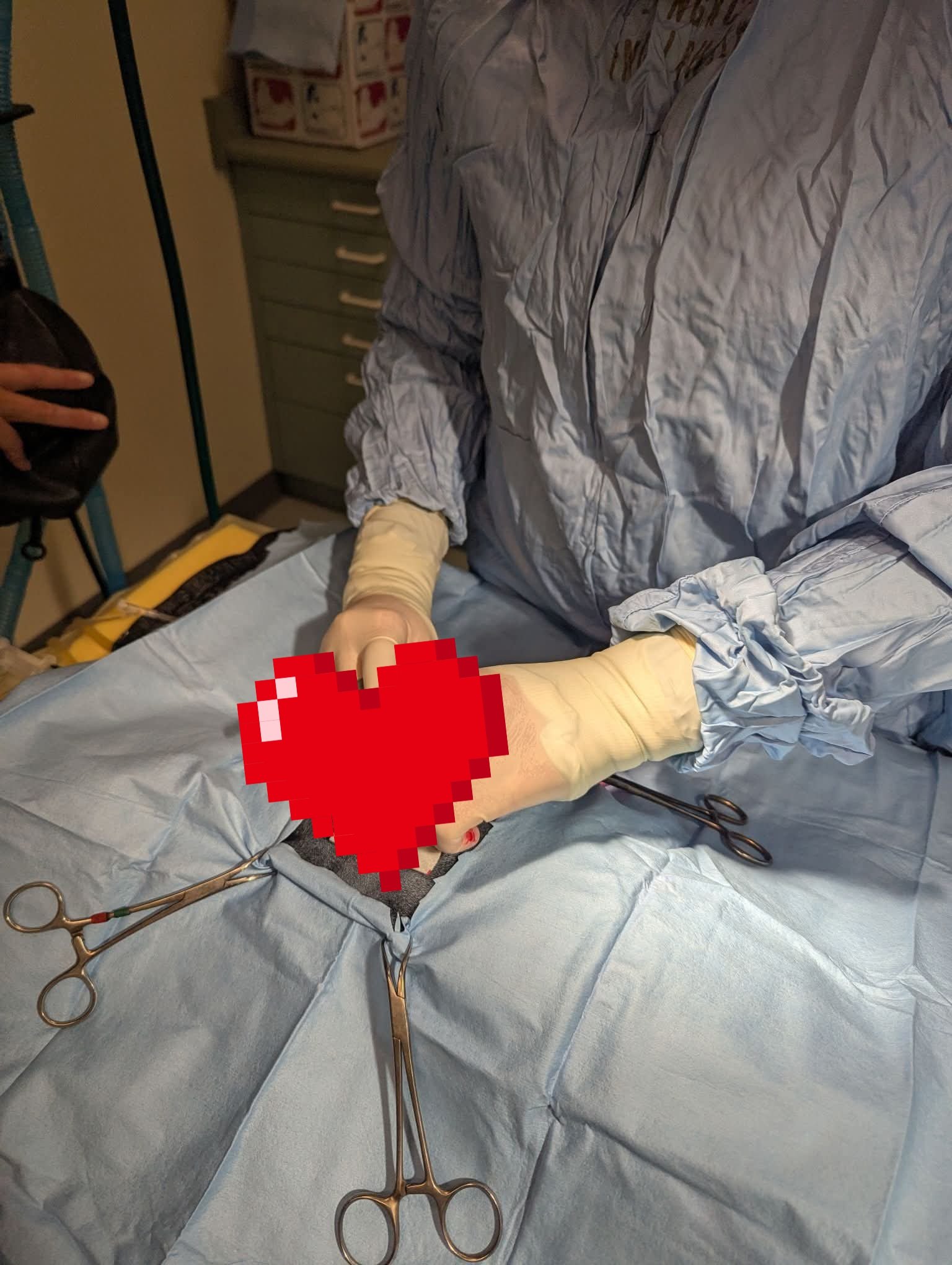 A medical procedure in a hospital room involving surgical tools like scissors on a table near a patient lying on a bed. A person wearing a gray protective gown and gloves is present, with a hand holding some medical equipment or tools. The patient’s face is pixelated for privacy.