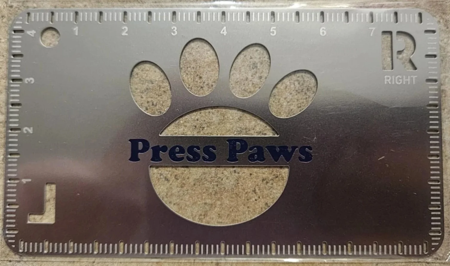 Metal ruler with paw print cutout and the words 'Press Paws' on a concrete surface.