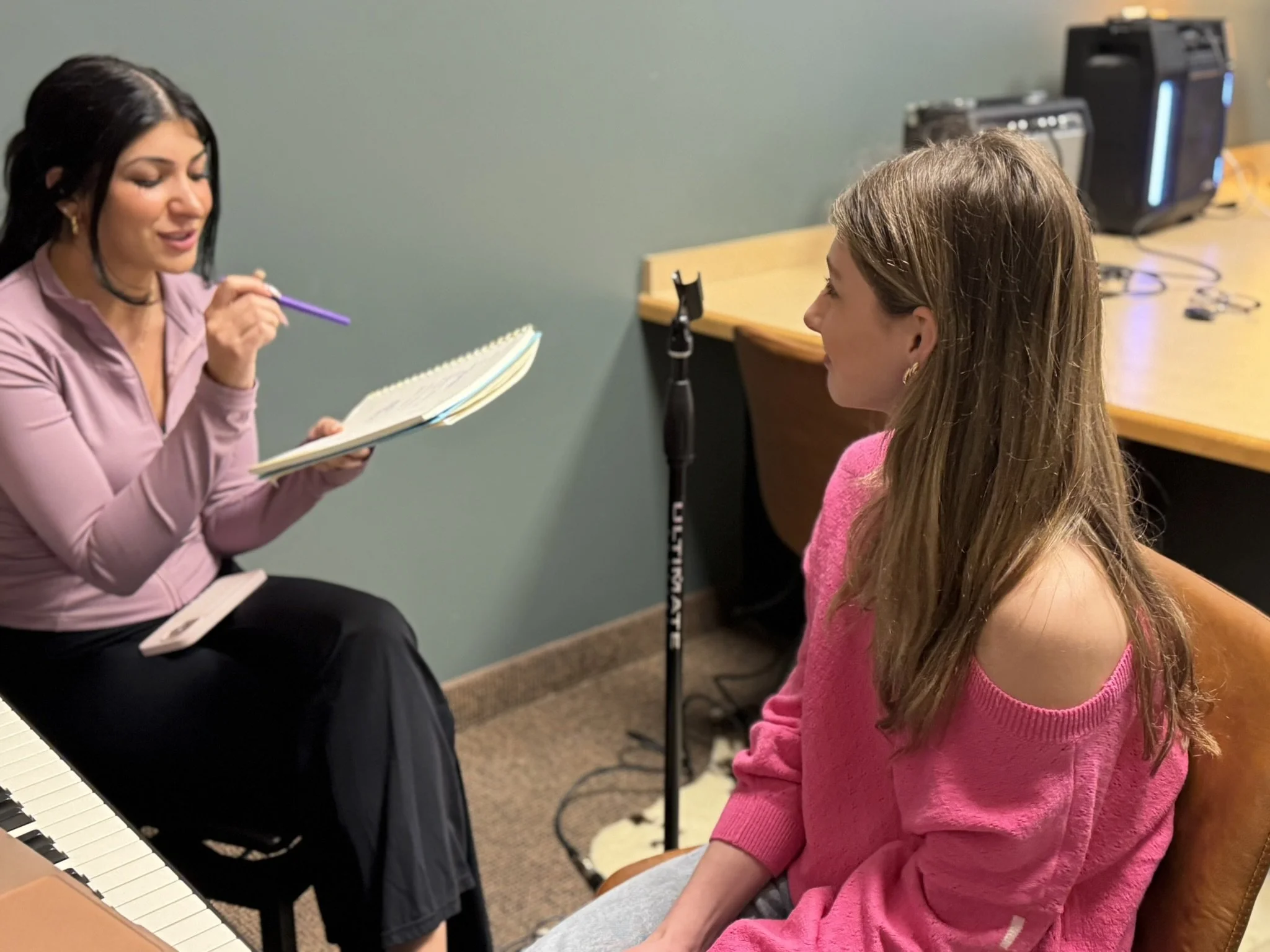 A young girl in a pink sweater sitting in front of a woman who is holding a notebook and a pen, appearing to be in a music or voice lesson setting with a keyboard and audio equipment in the background.